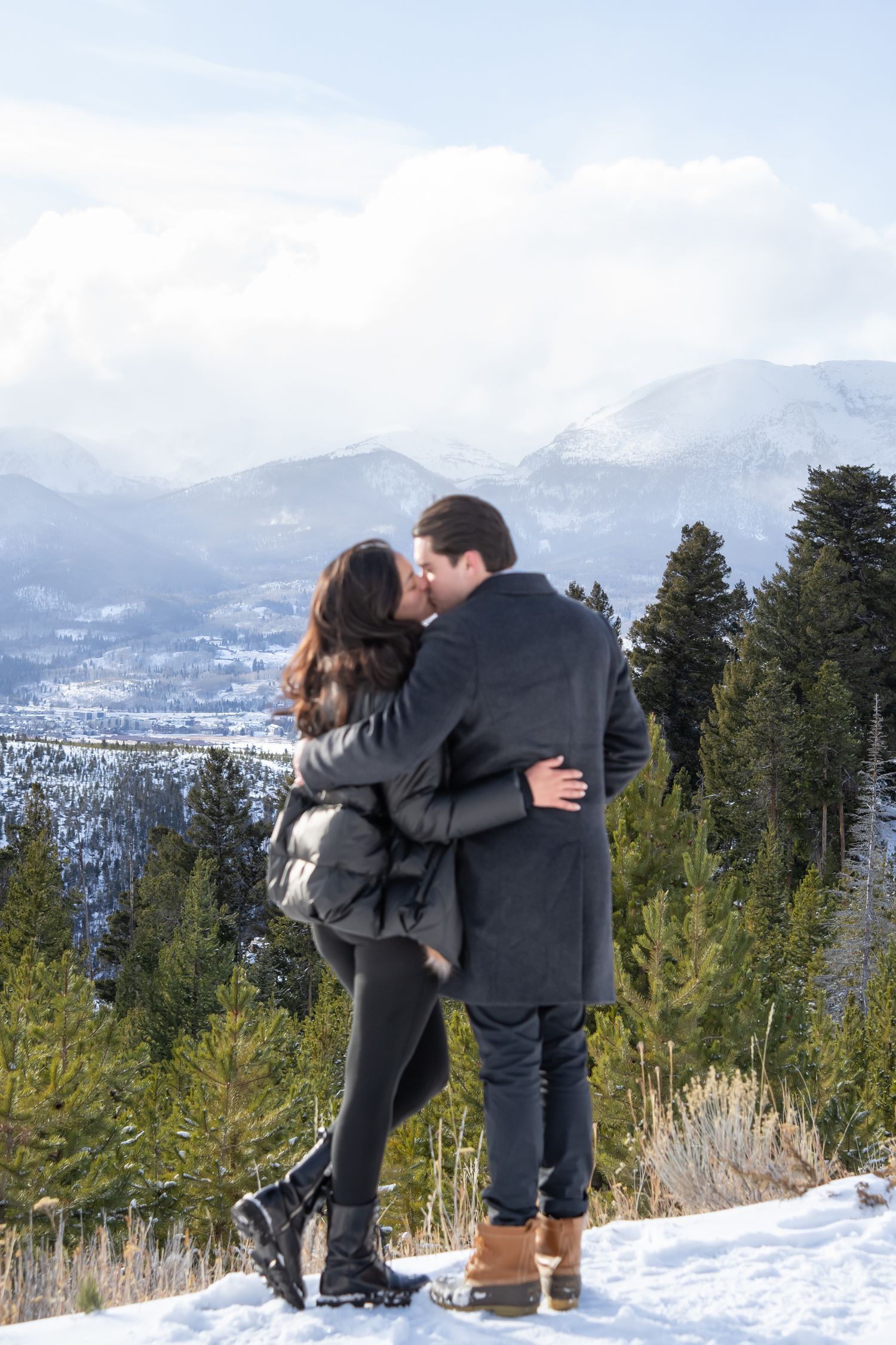 A couple embraces while enjoying a scenic winter mountain view with snow-covered peaks and evergreen trees in the background.