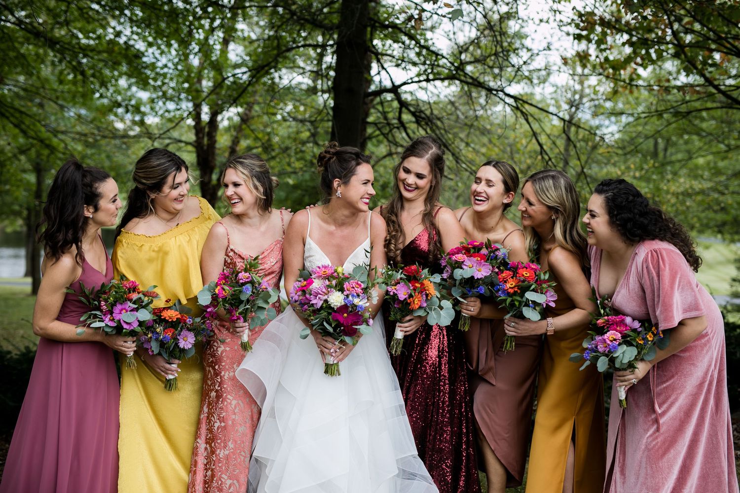 A wedding party in pink, yellow, and burgundy dresses poses together outdoors surrounded by lush greenery.