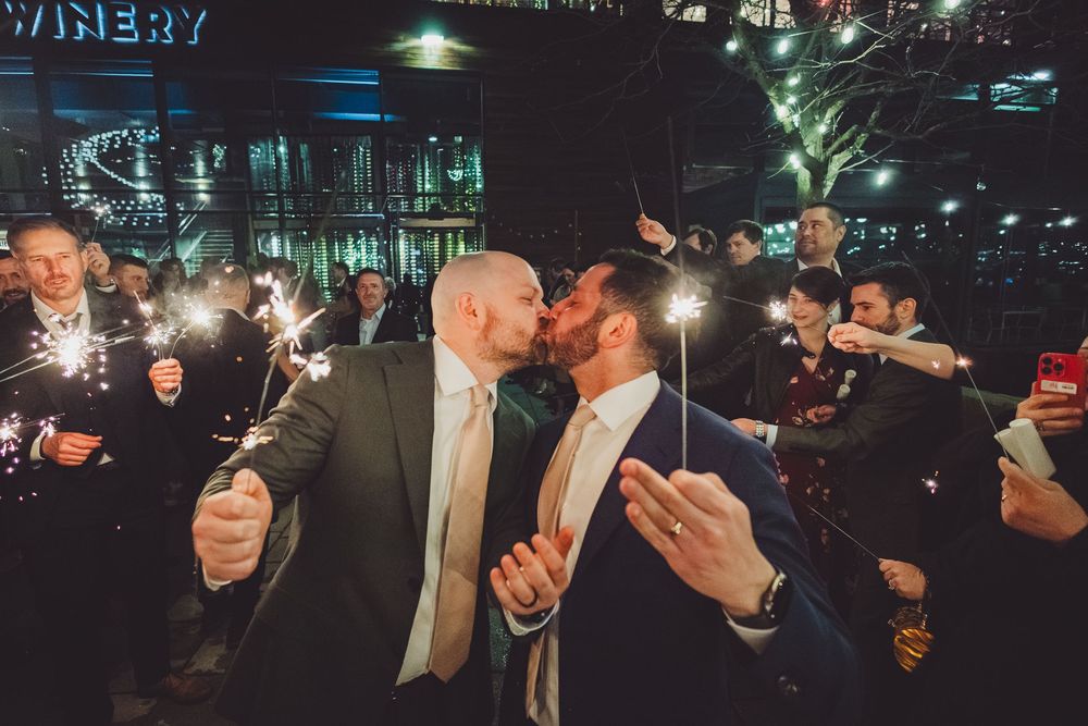 A married gay couples shares a romantic kiss during their wedding exit celebration at night surrounded by guests holding sparklers.