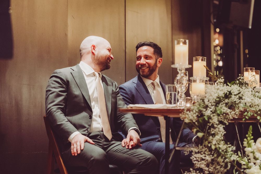 Two grooms in suits share a laugh while sitting at a table decorated with floral arrangements at an indoor venue.