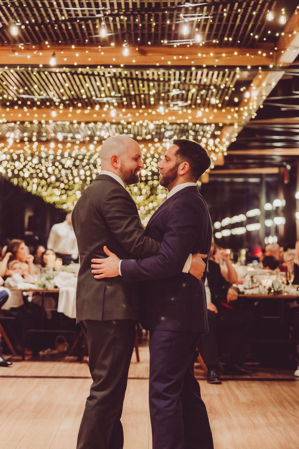 Married same-sex couple dancing under romantic string lights at an indoor reception venue with wooden beams and warm lighting.