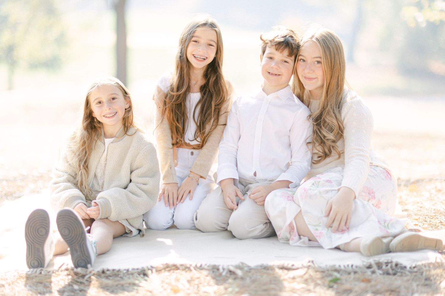 Children in light colored clothing sitting together outdoors on a white blanket in soft natural lighting.