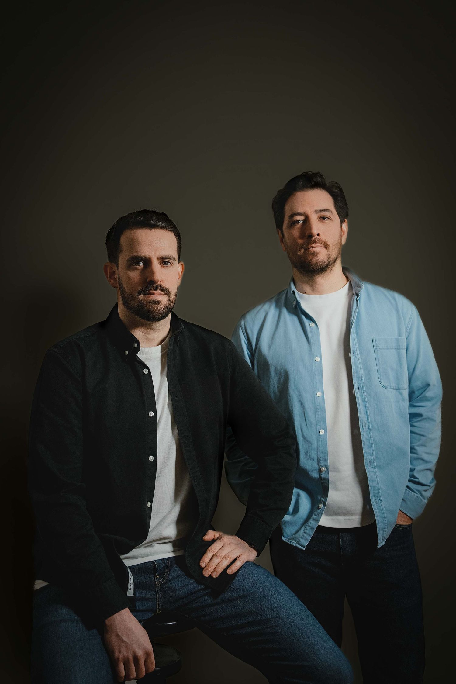 Two people in business casual attire sit casually on modern office furniture against a white wall.