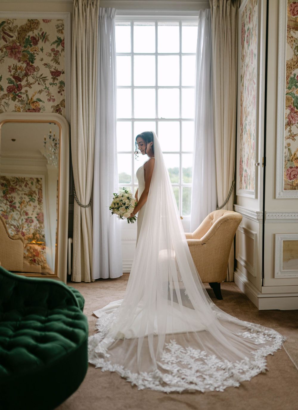 A flowing white wedding dress with a long lace-trimmed cathedral veil displayed in an elegant room with floral wallpaper.
