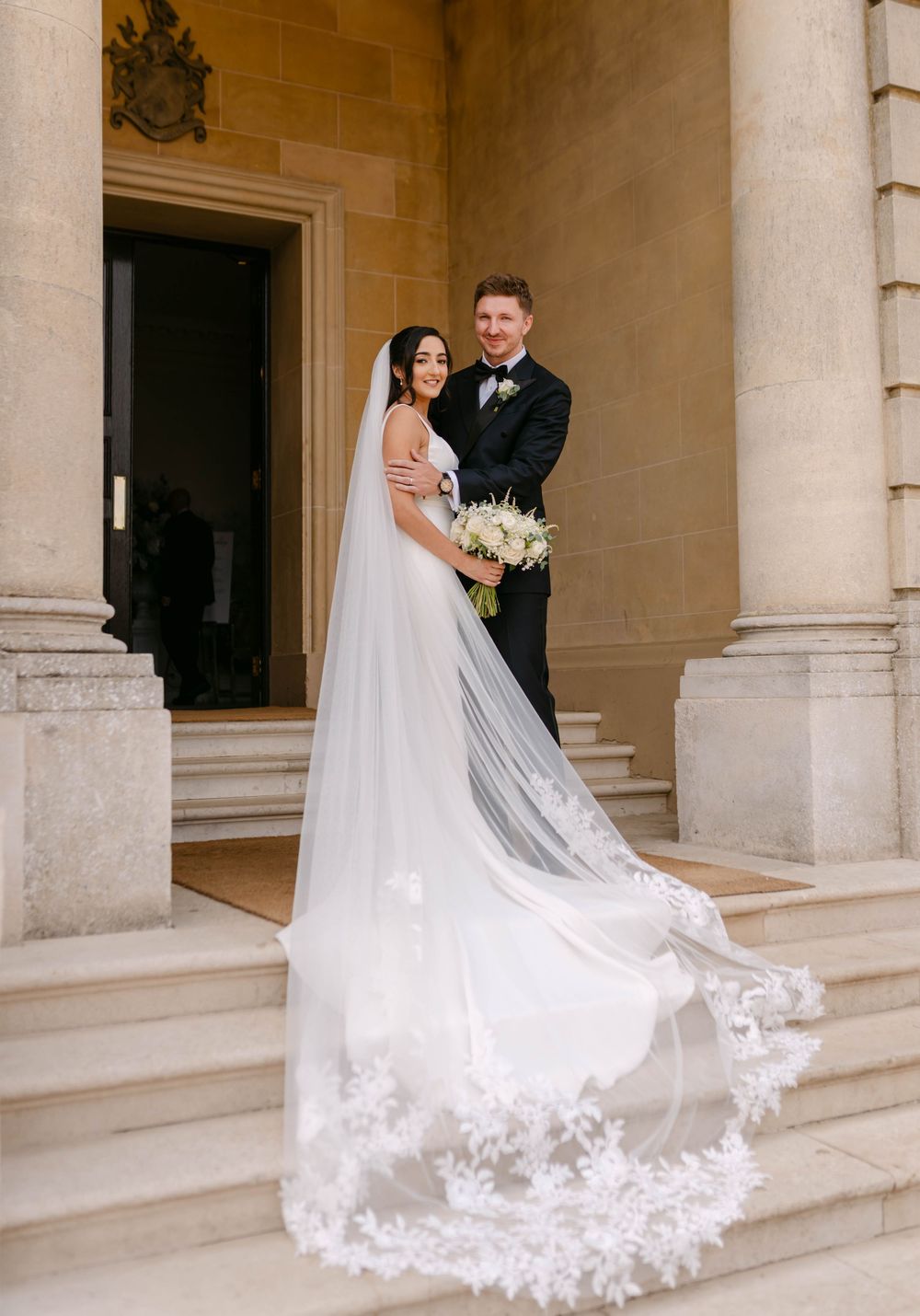 Elegant wedding couple poses on stone steps with a flowing lace-trimmed veil trailing behind the bride.