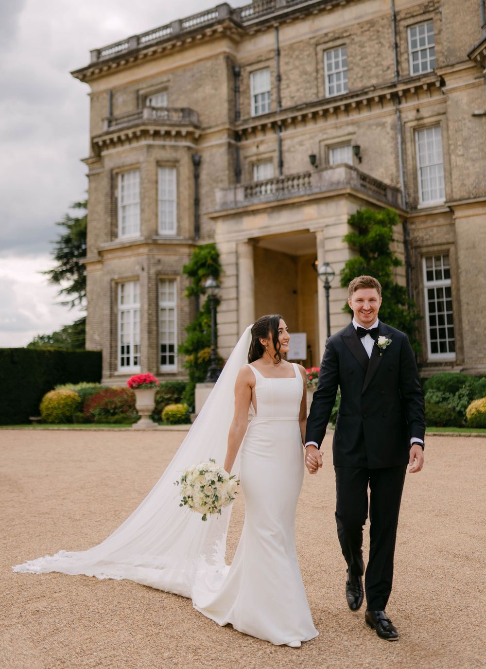 A wedding couple walks together on a gravel path in front of a grand historic mansion with elegant stone architecture.