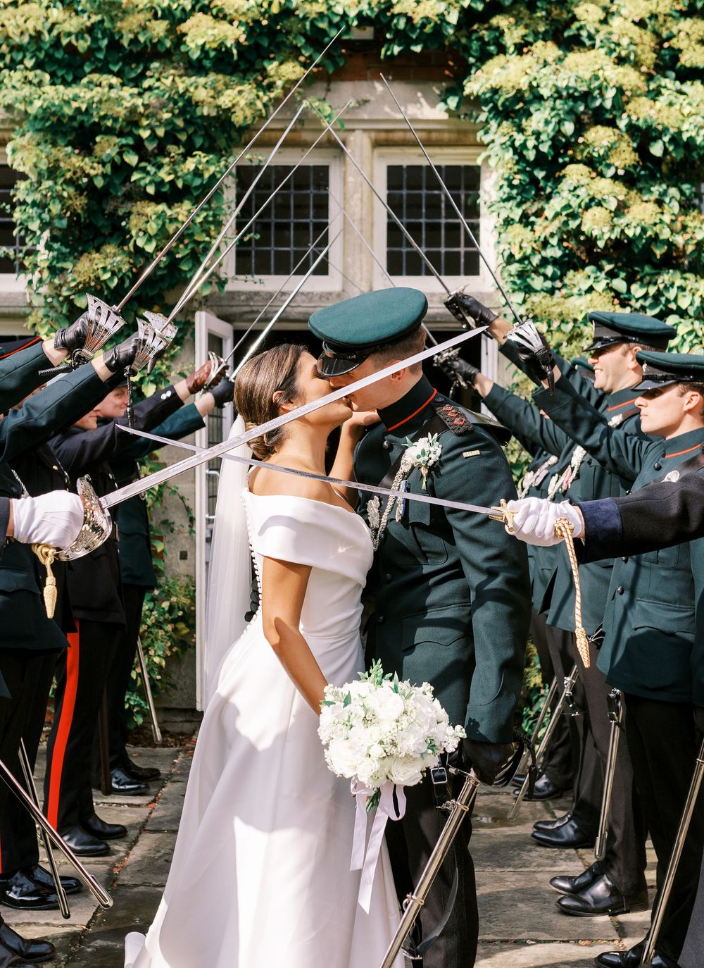 Military officers perform a ceremonial sword arch at an outdoor wedding ceremony with white flowers and greenery.