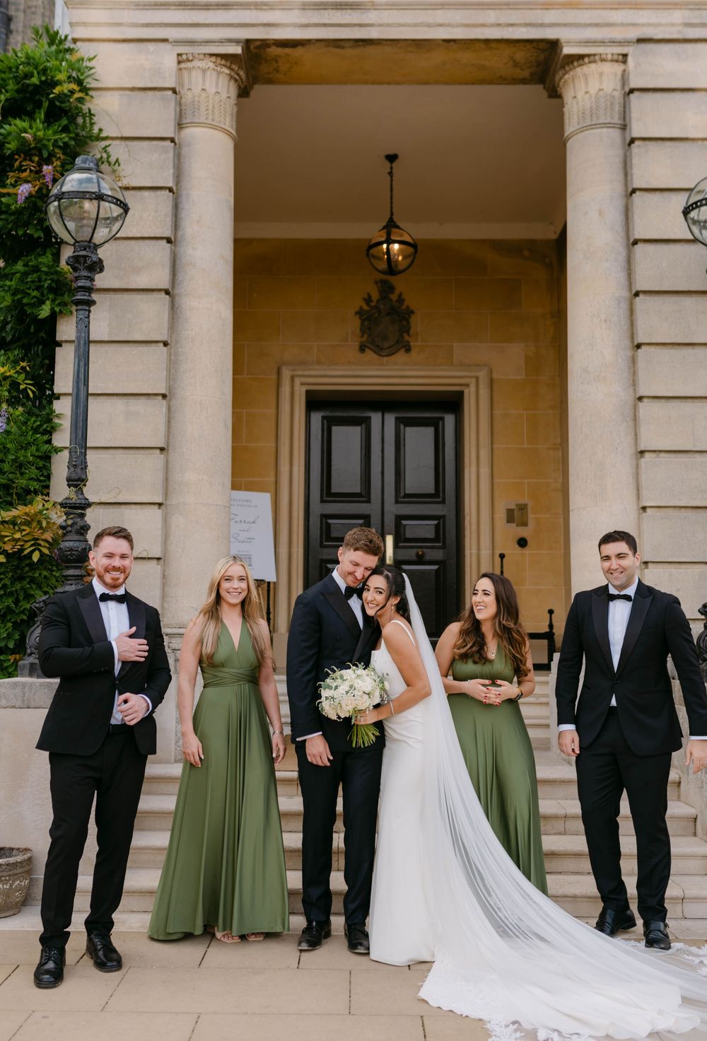 Wedding party poses on elegant stone entrance steps with sage green bridesmaid dresses and classic black tuxedos.