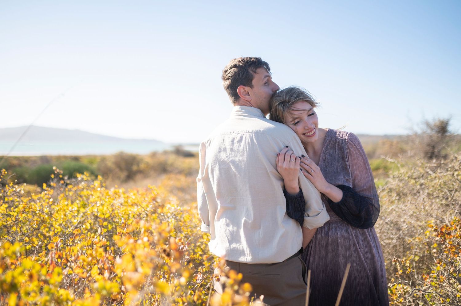 Couple embracing in a sunny wildflower field, man kissing woman's cheek outdoors.
