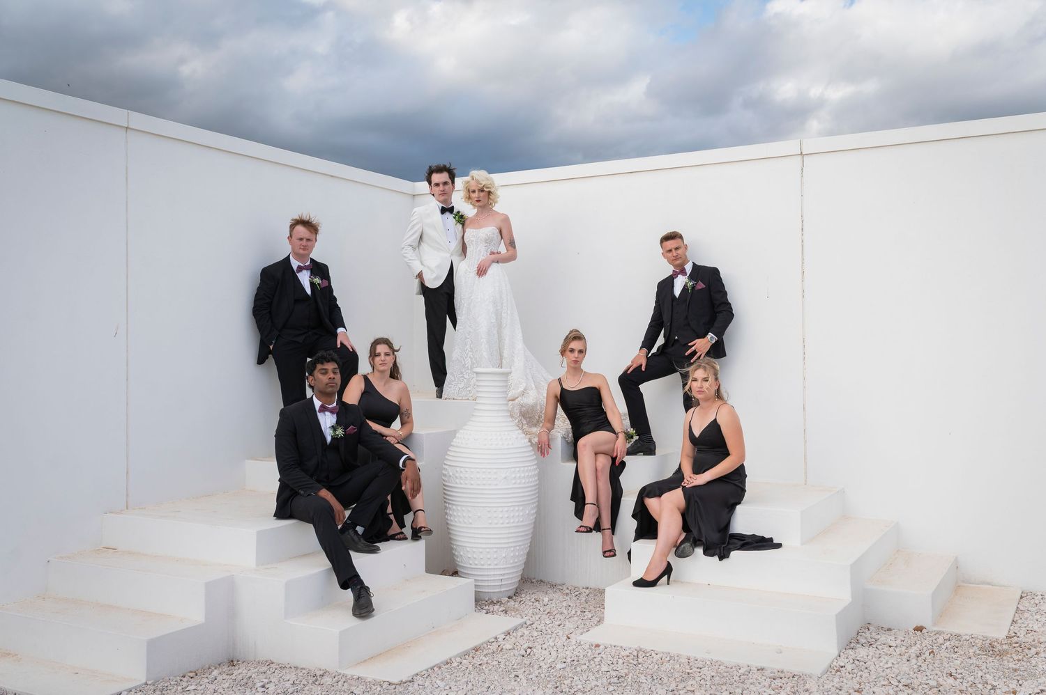 Wedding party in black and white attire posed on white steps with a large vase outdoors under cloudy skies.