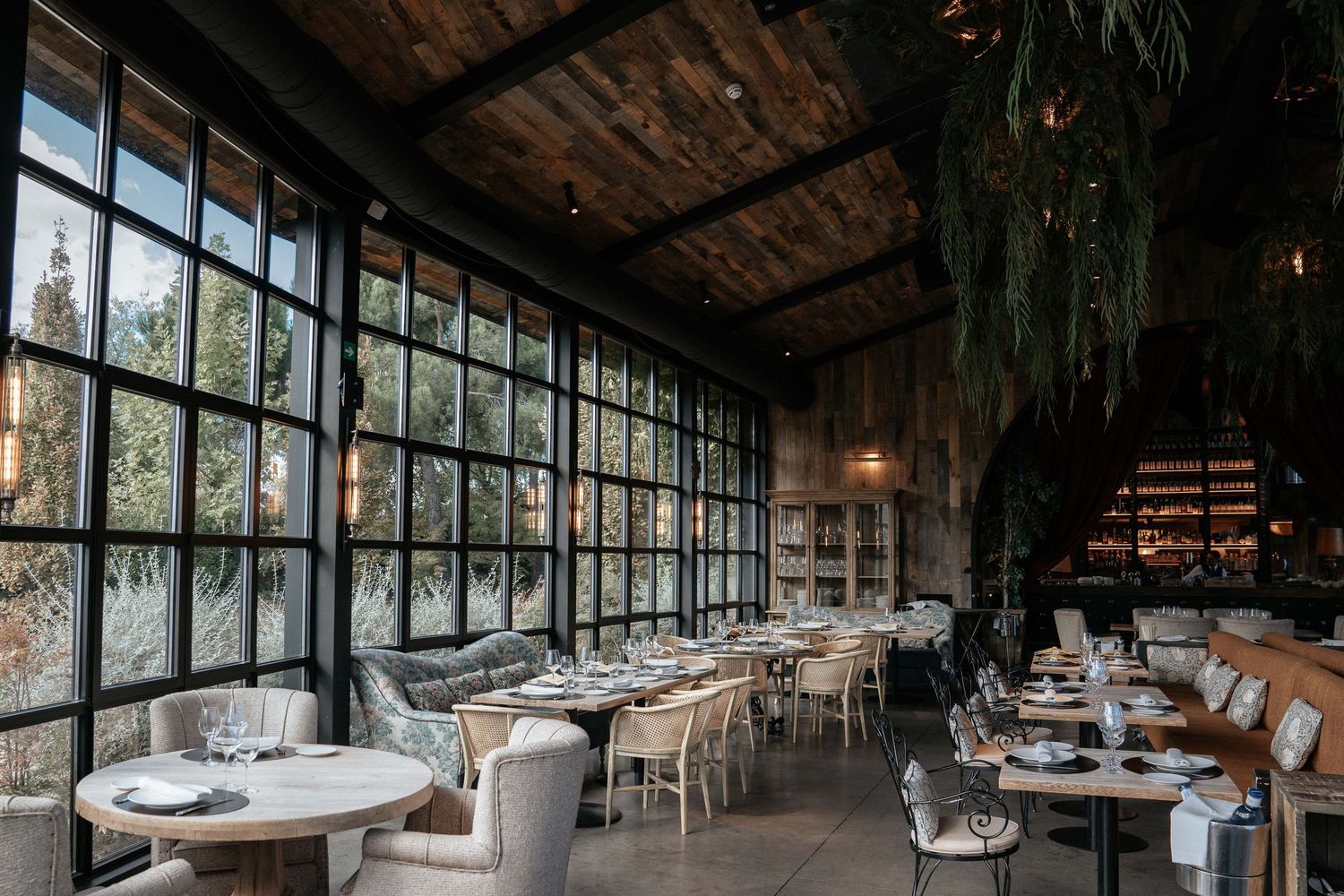 Modern rustic restaurant interior with floor-to-ceiling windows, wooden beams, and elegant table settings.