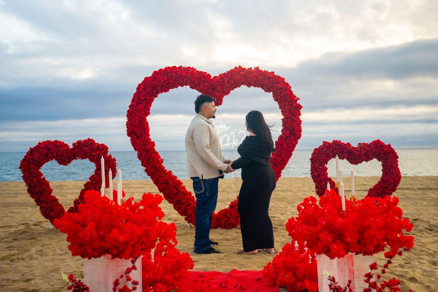 A romantic beach proposal setup with large red heart-shaped flower arches and rose petals on the sand at sunset.