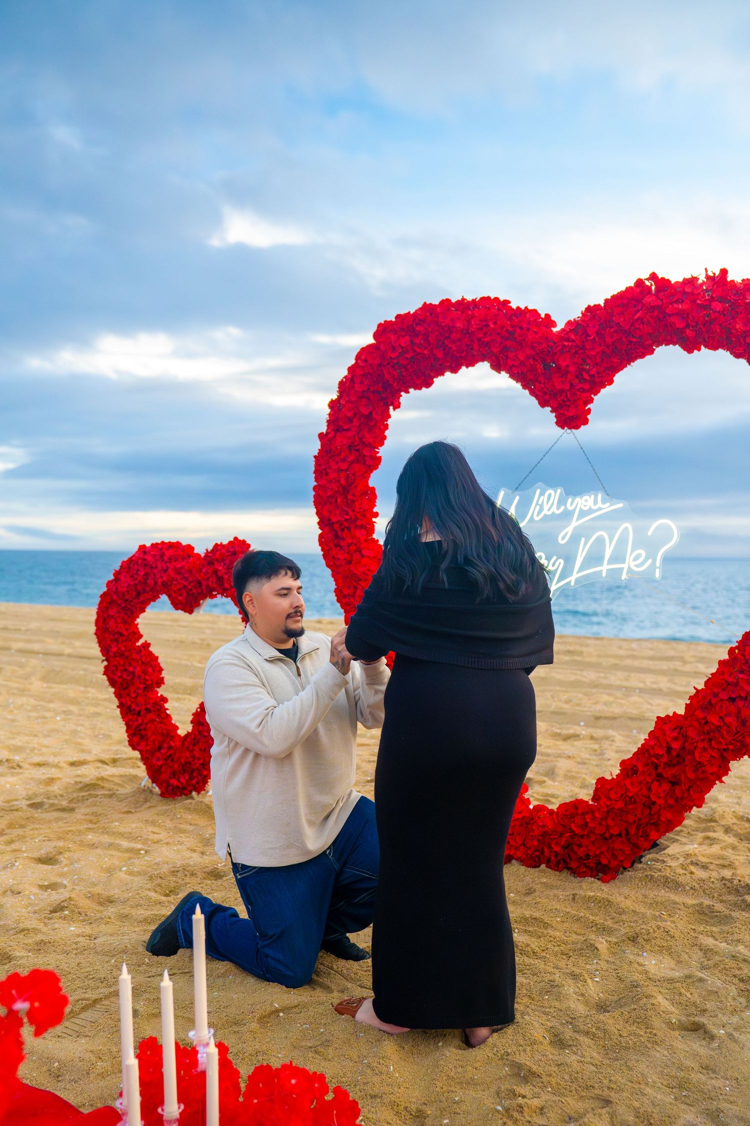 A romantic beach proposal setup with red heart-shaped flower arrangements and candles on the sand at sunset.