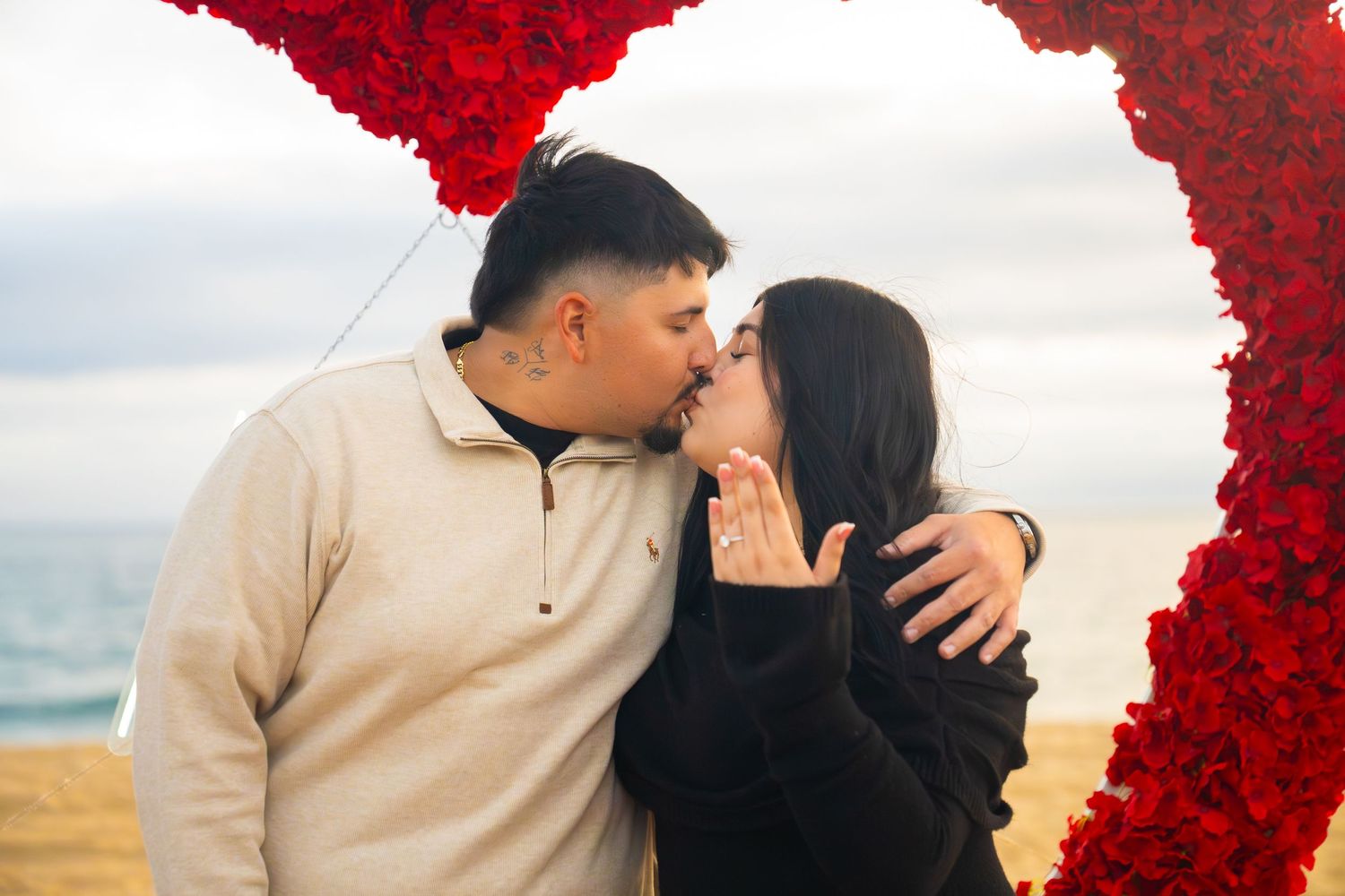 A romantic couple embraces near a vibrant red floral arch at sunset during a beach photoshoot.