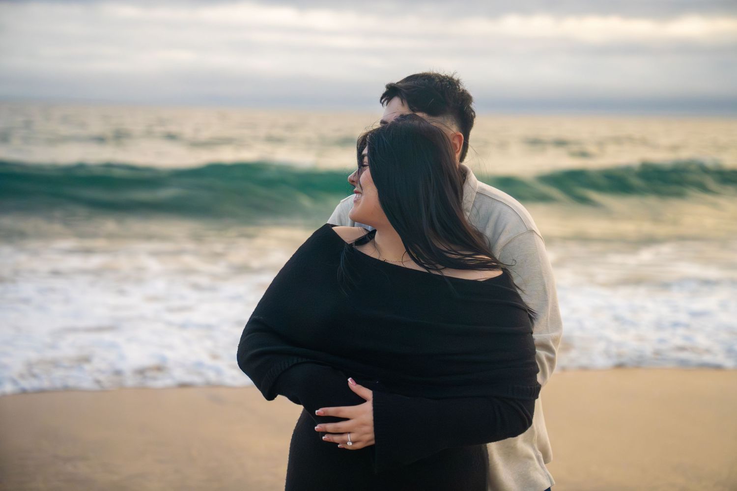 A couple embraces on a sandy beach at sunset with ocean waves in the background.