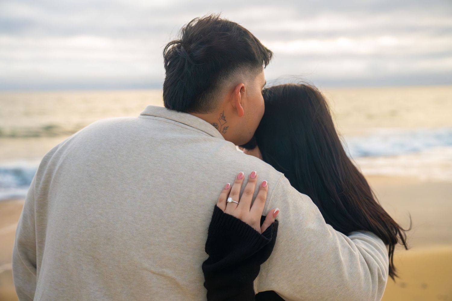 A couple embraces romantically on a sandy beach at sunset with waves in the background.