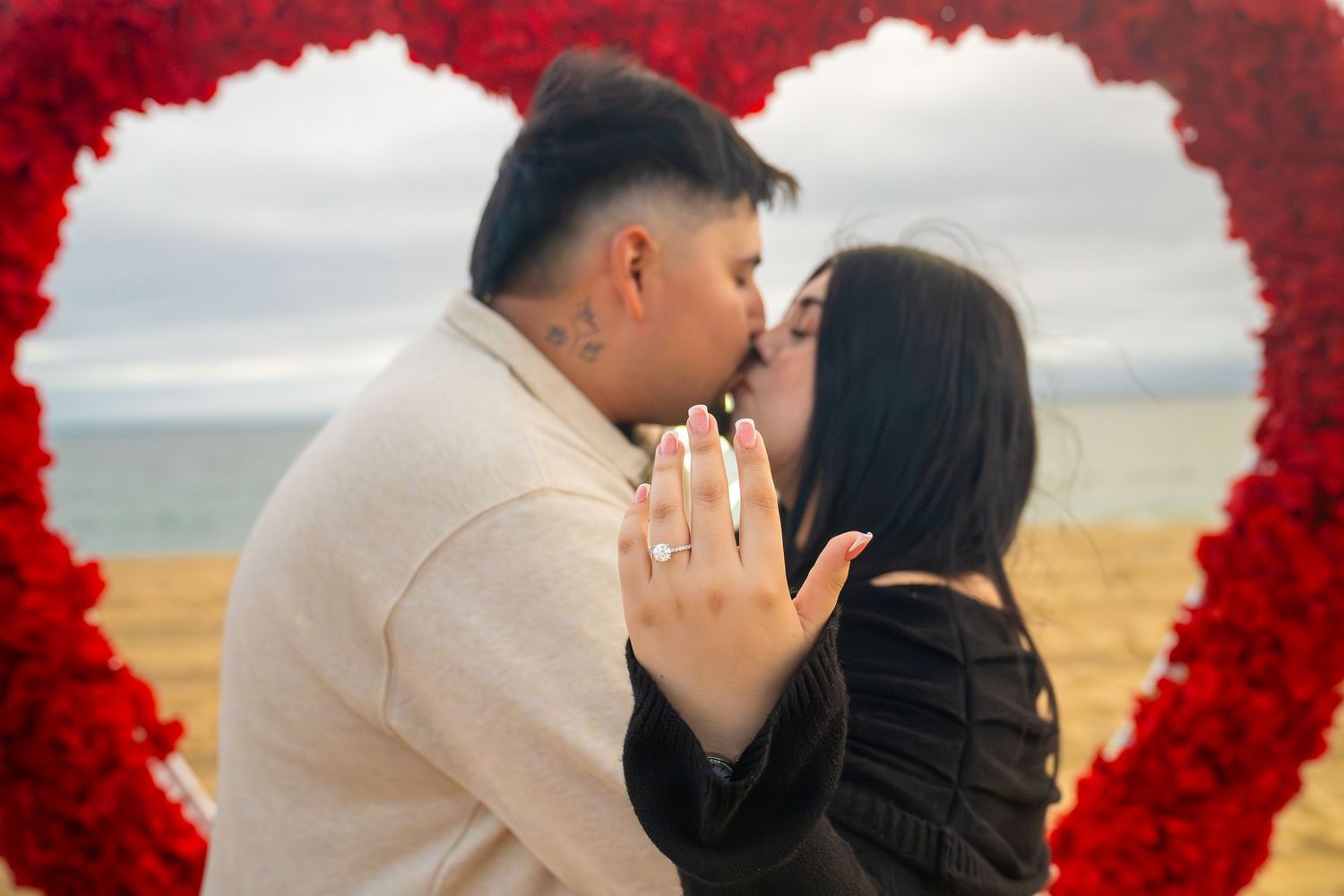 A romantic couple shares a tender kiss framed by a heart-shaped arch of red roses against a scenic coastal backdrop.