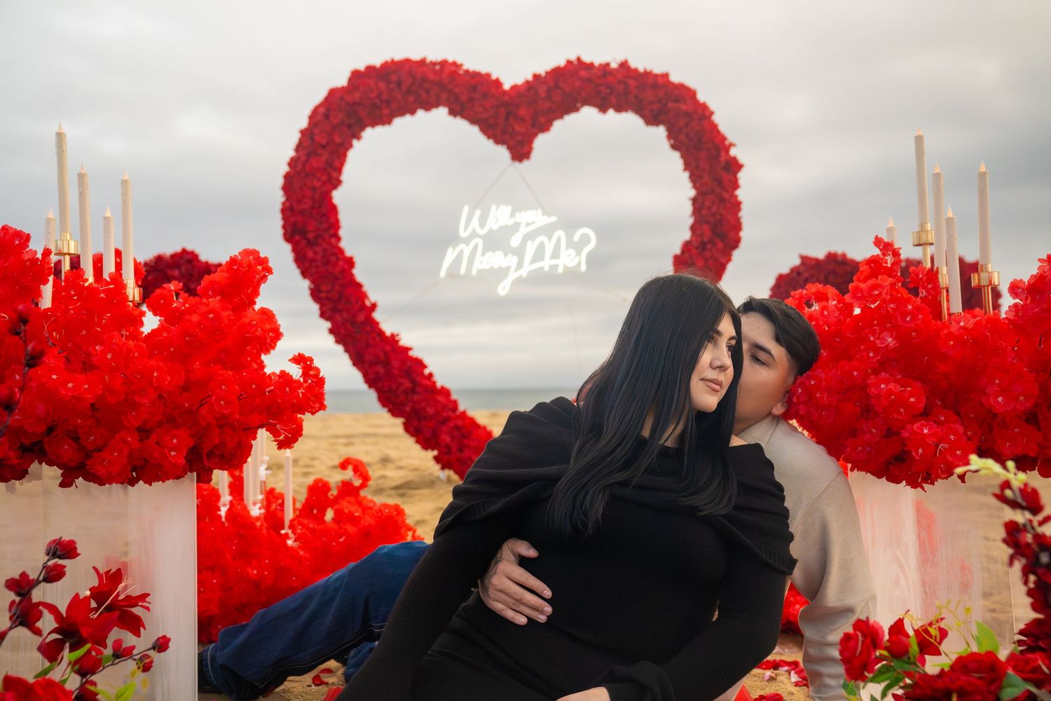 A couple embraces in front of vibrant red heart-shaped decorations at an outdoor location.