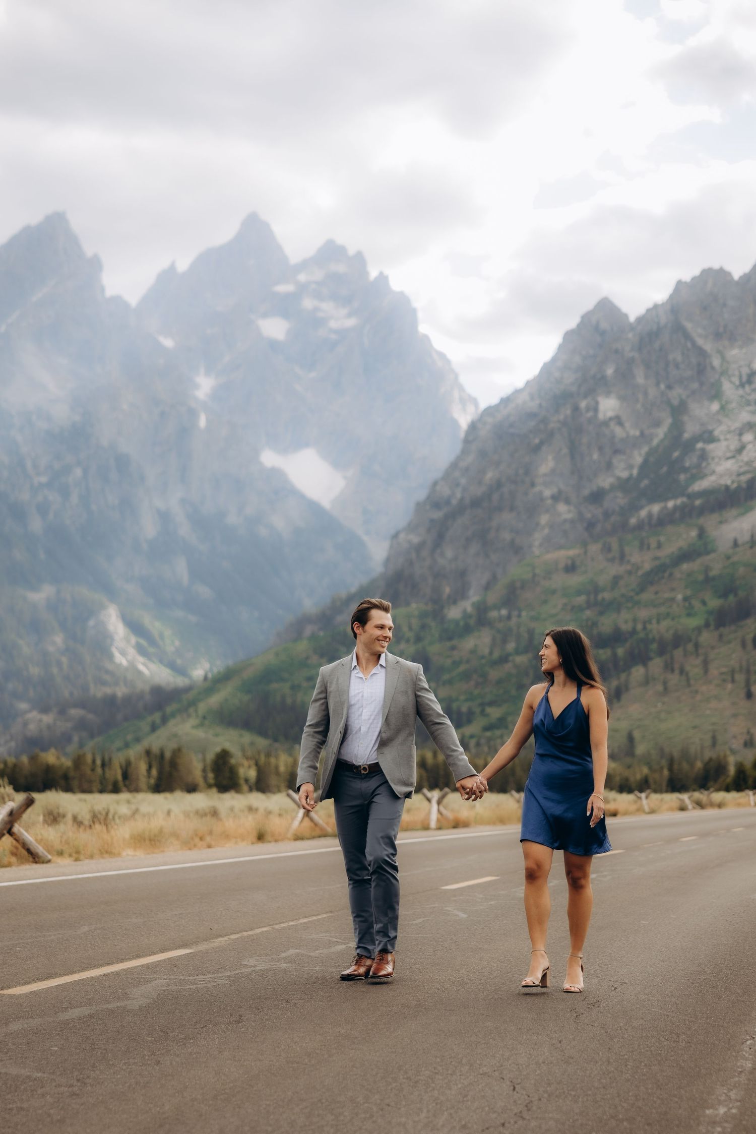 Two people walk together along a scenic mountain road with dramatic peaks rising in the background at Grand Teton National Park.