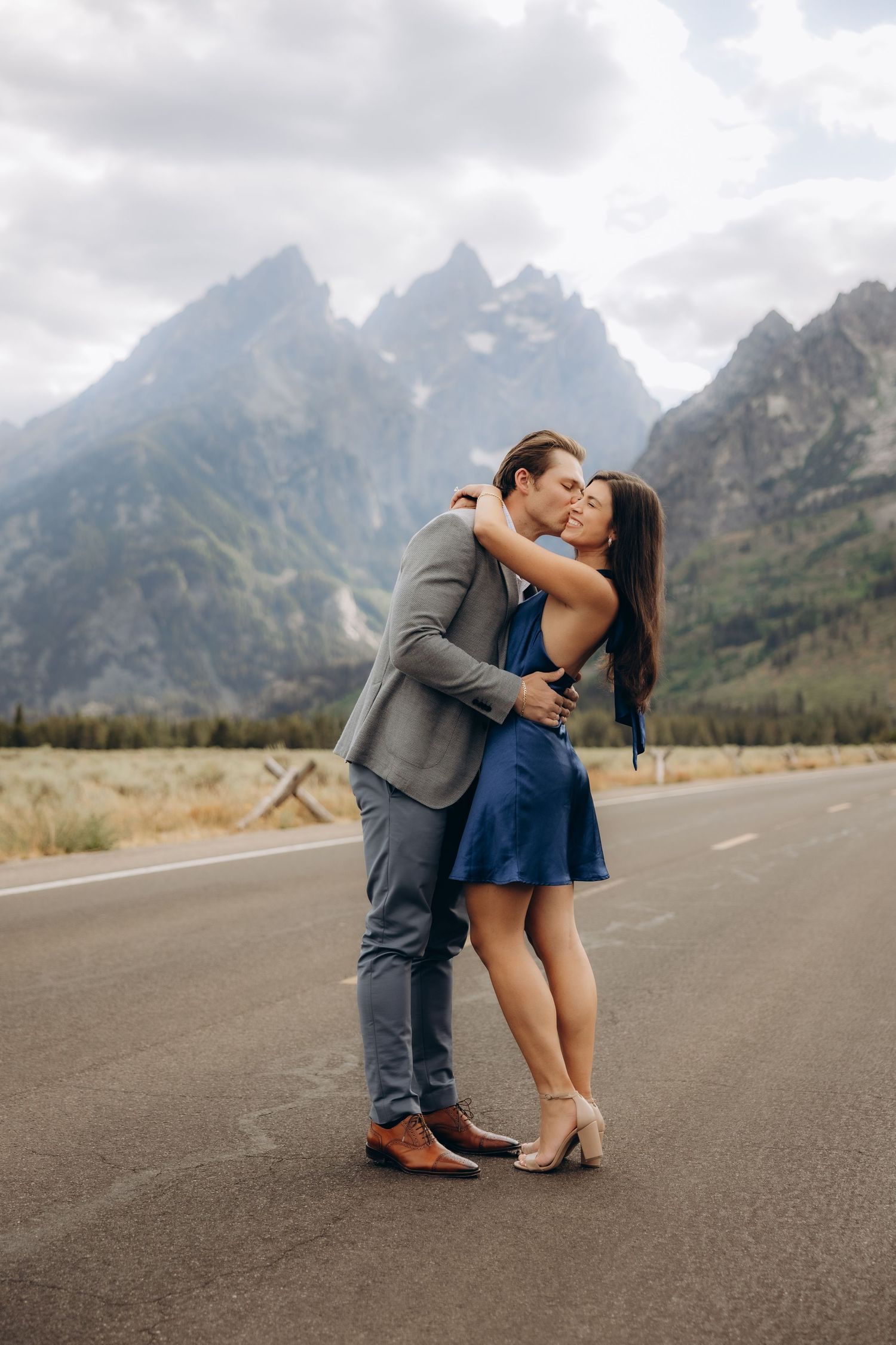 Couple shares intimate moment on mountain road with Grand Tetons in background.