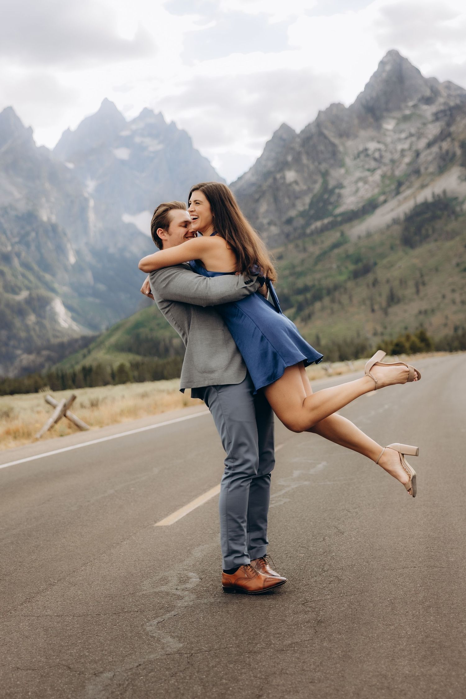 A couple shares a romantic spinning embrace on an empty mountain road with dramatic peaks in the background.