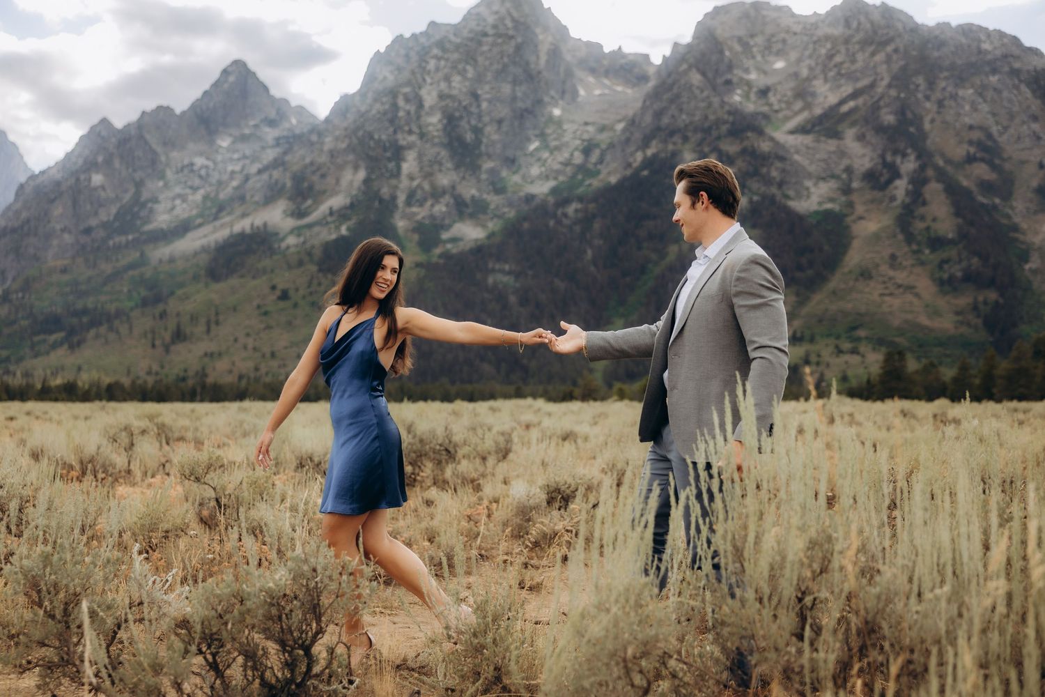 Two people hold hands while standing in a grassy meadow with dramatic Grand Teton mountains rising in the background.