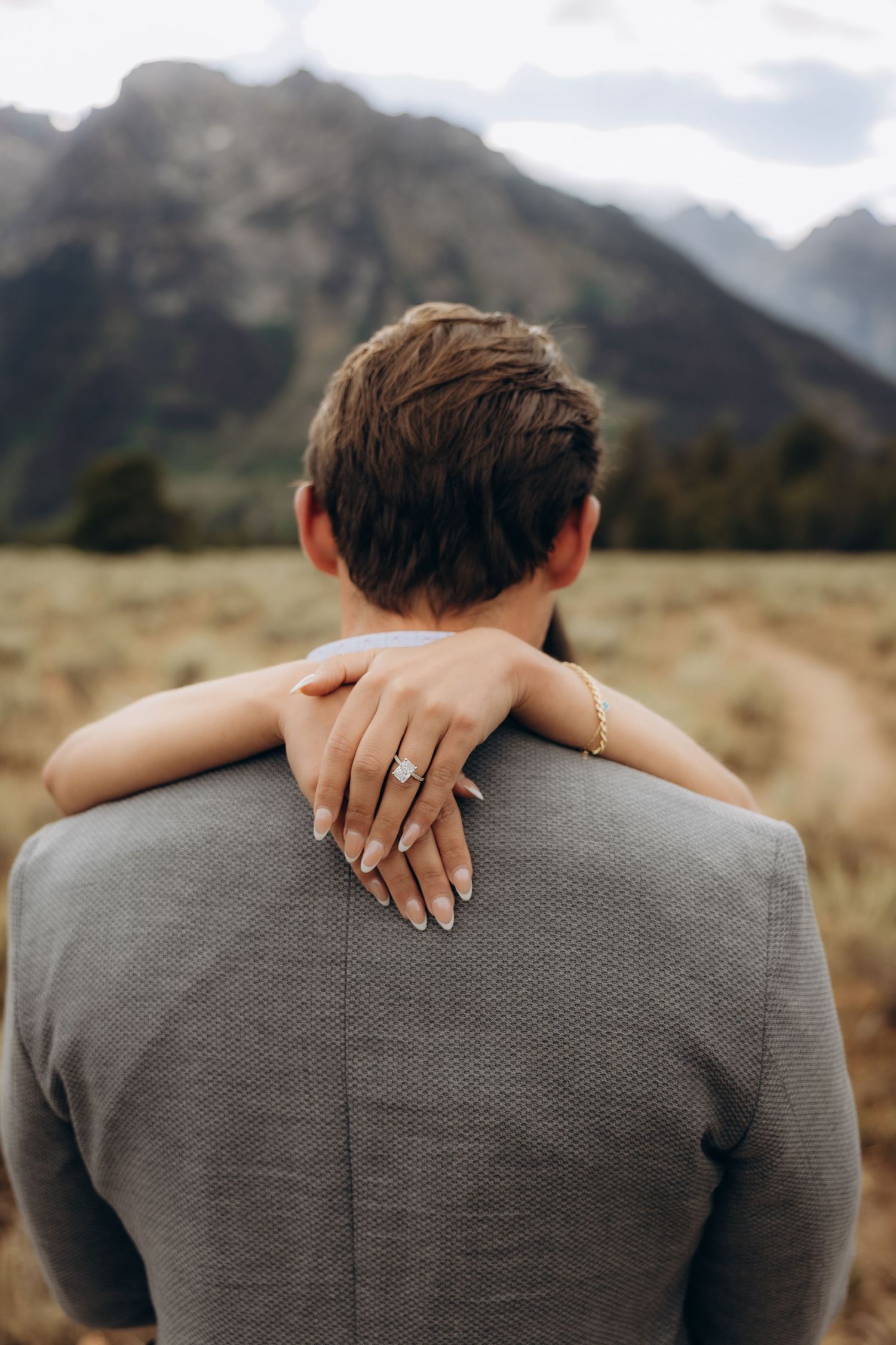 Intimate moment between couple with mountain backdrop as bride embraces groom from behind in grey suit.