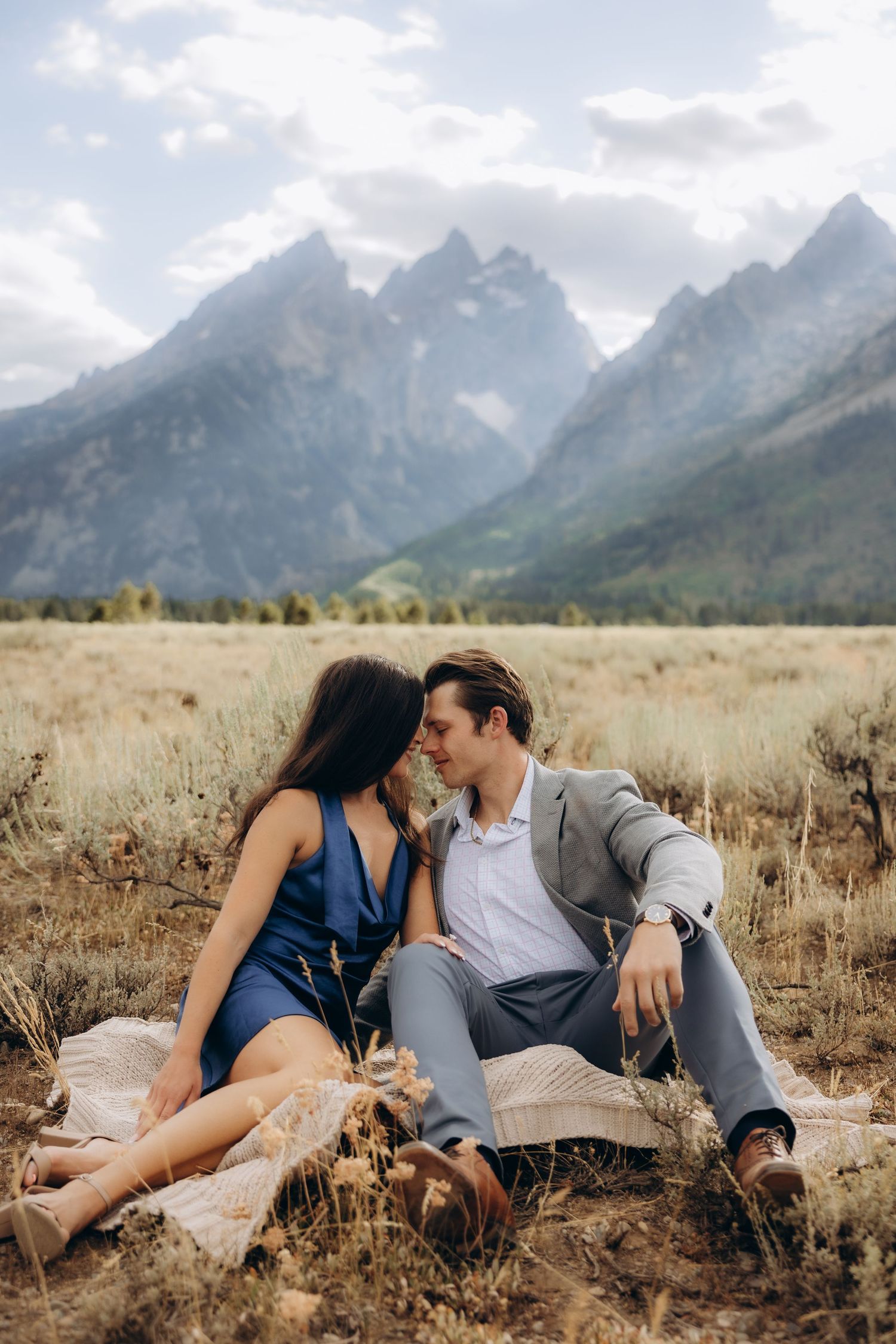 Couple lying together in a grassy field with majestic mountain peaks rising in the background under cloudy skies.