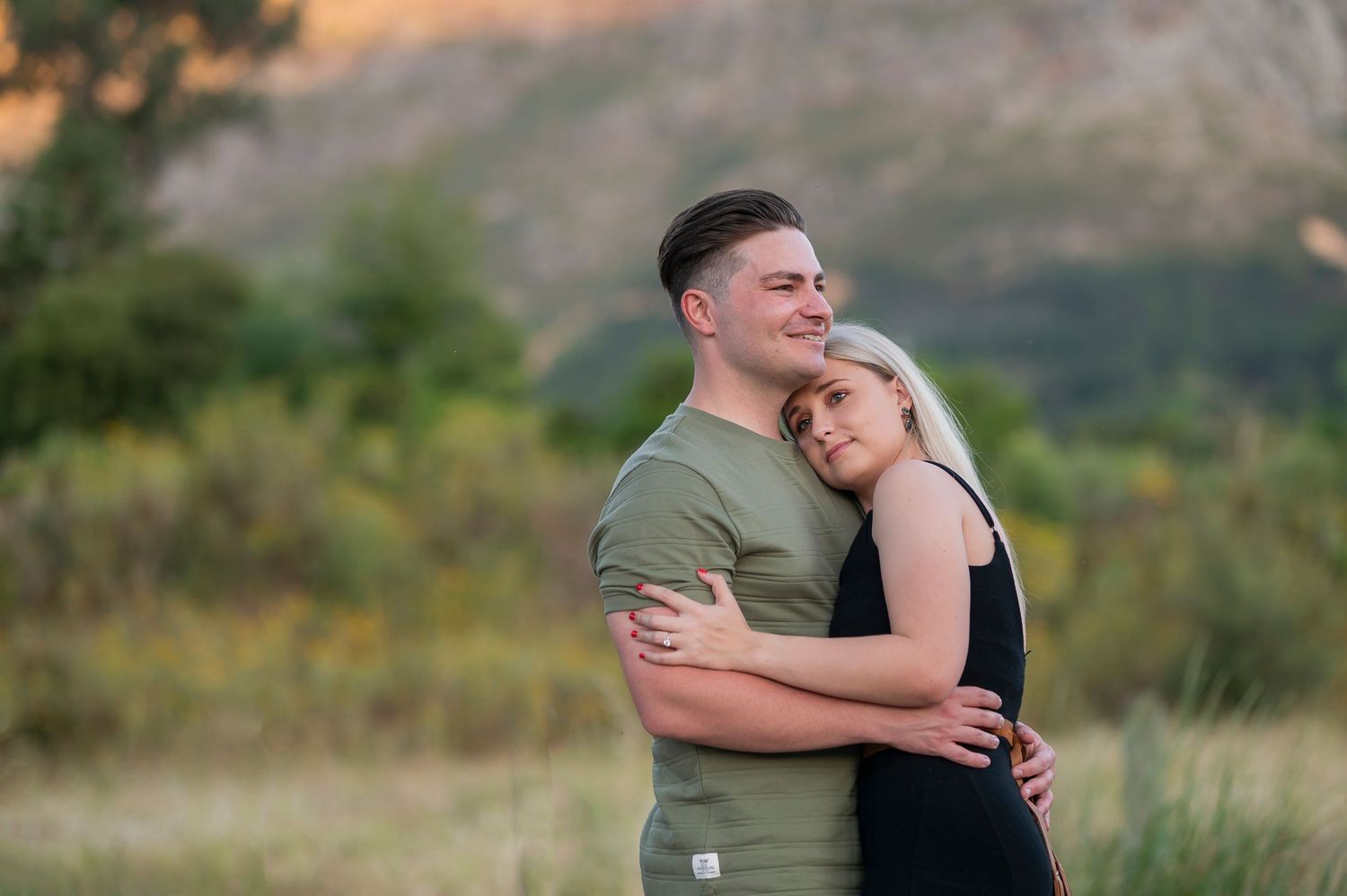 A couple embraces tenderly outdoors against a soft mountain landscape background during golden hour.