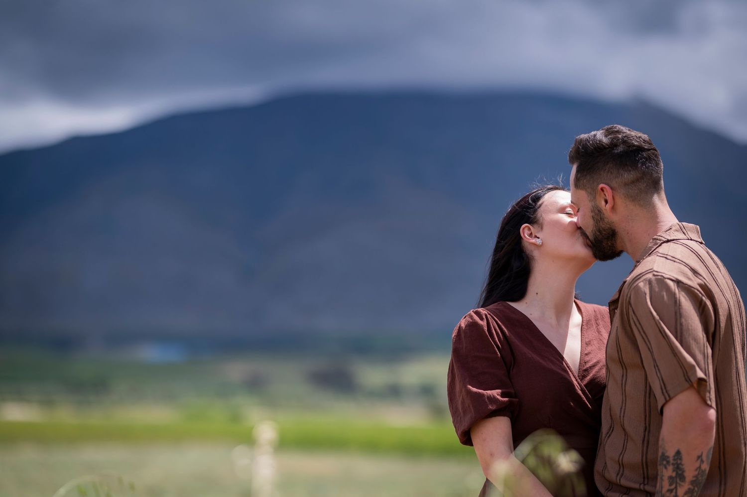 A romantic couple embraces in front of mountains with green hills and dark clouds in the background.