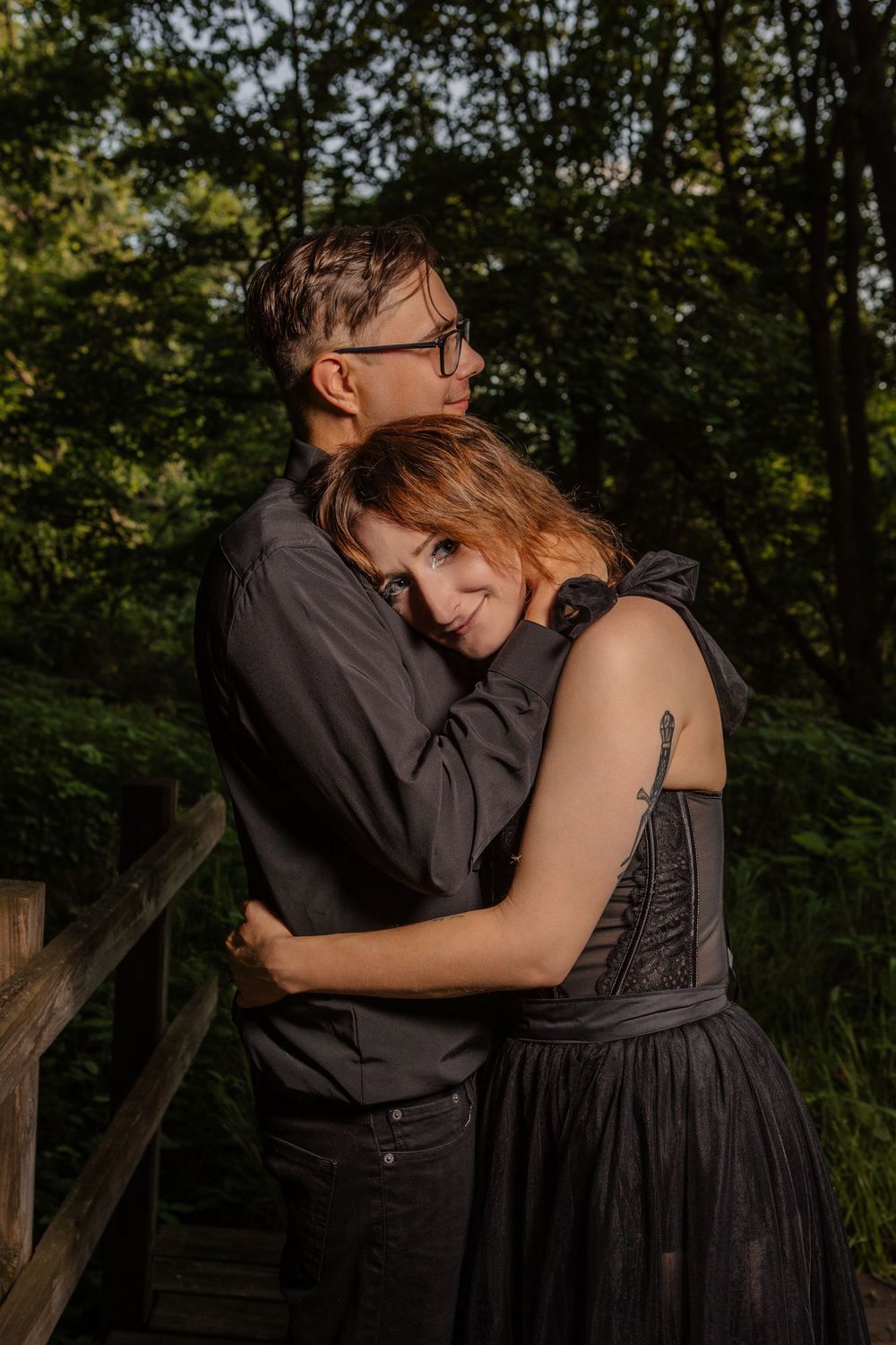 A couple embraces on a rustic wooden walkway surrounded by green foliage.
