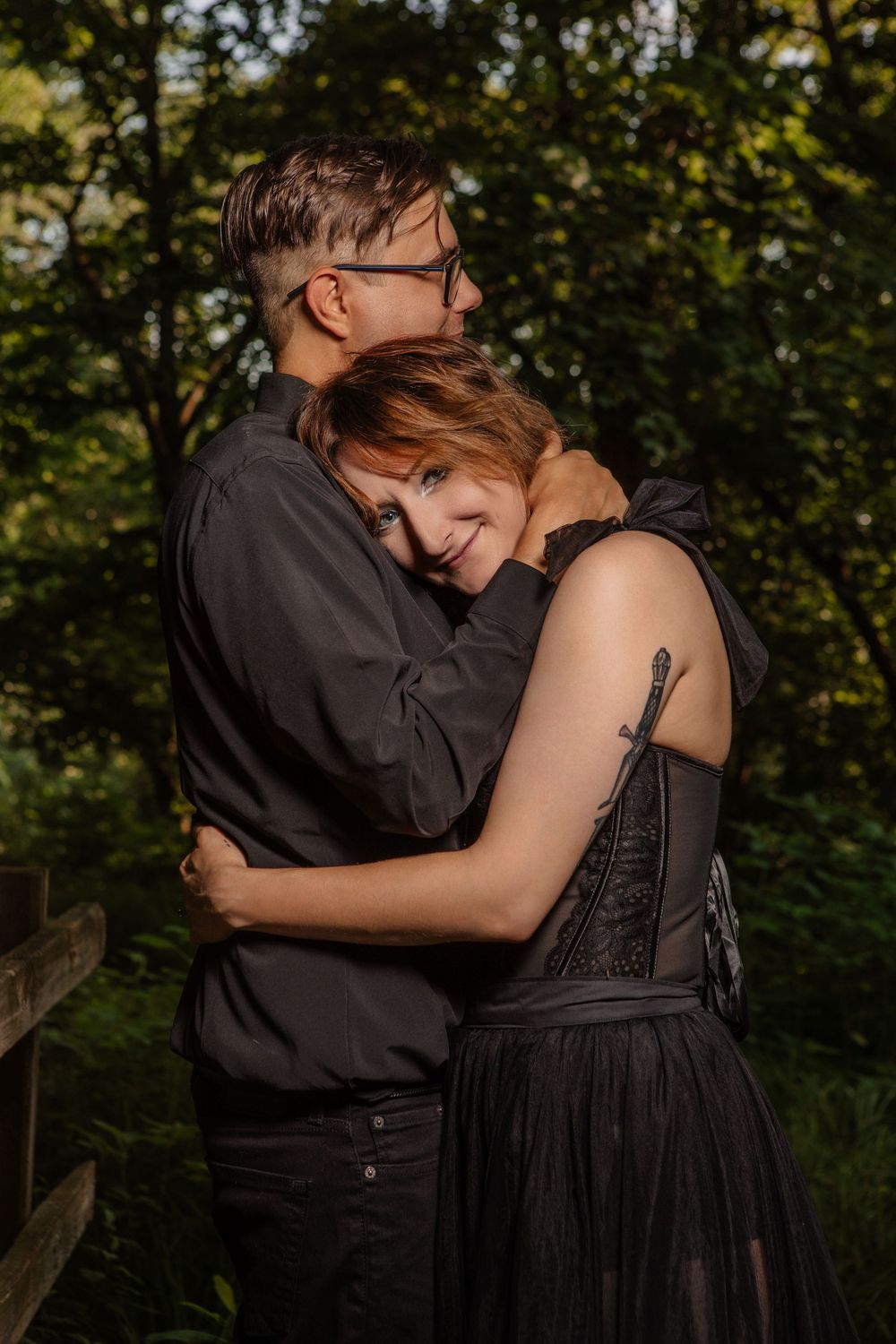 A couple in black attire embraces tenderly in a wooded setting during golden hour.