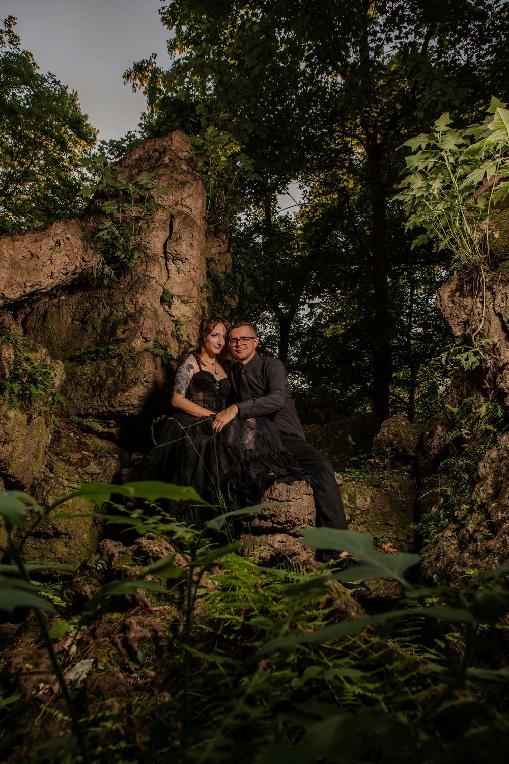 A couple poses intimately against rocky formations in a wooded natural setting.