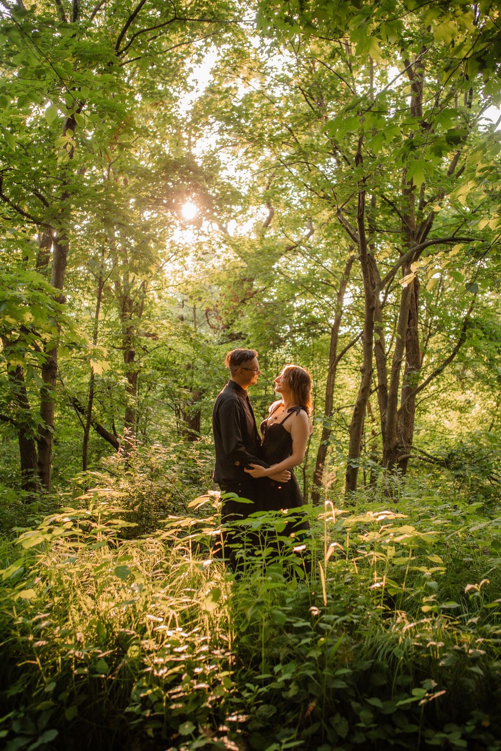 A couple stands together in a serene forest setting with sunlight streaming through trees.