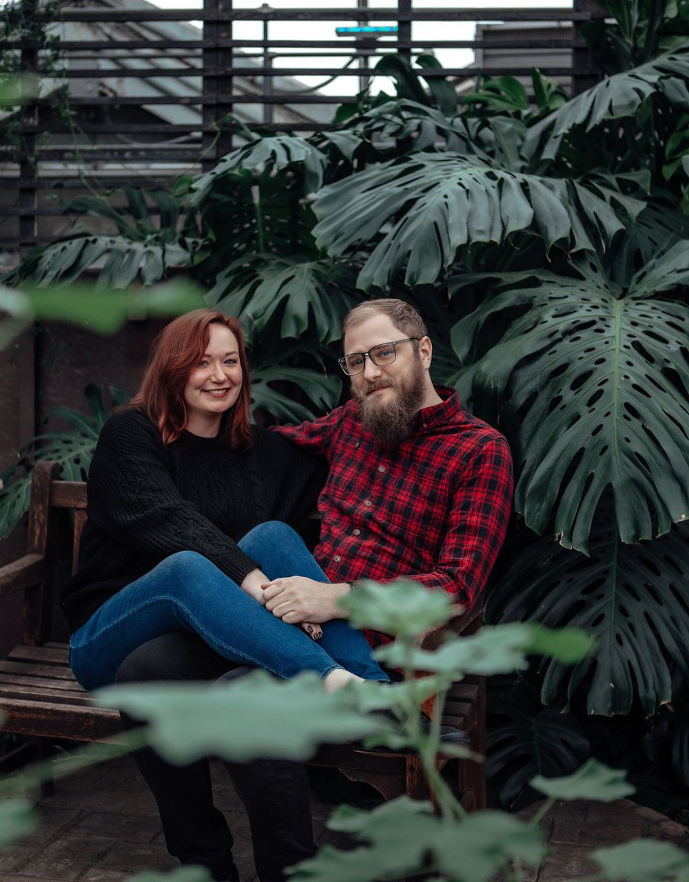 A couple sits together on a bench surrounded by large tropical monstera leaves in an indoor garden setting.