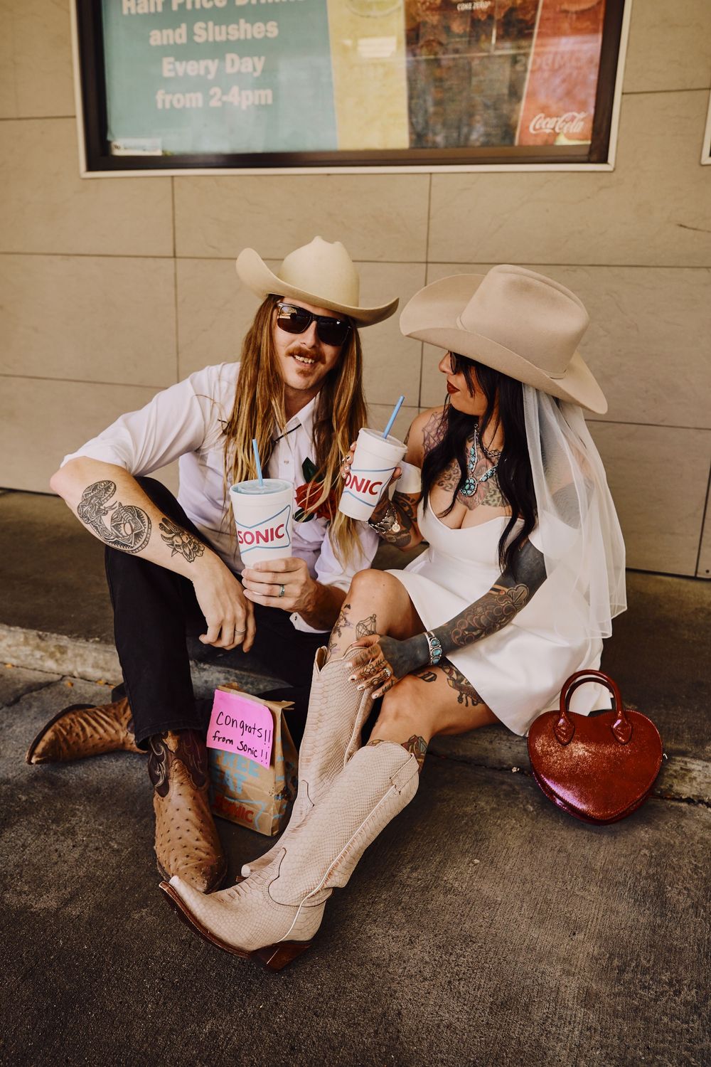 Two friends sitting on ground sharing drinks and laughs in casual setting.