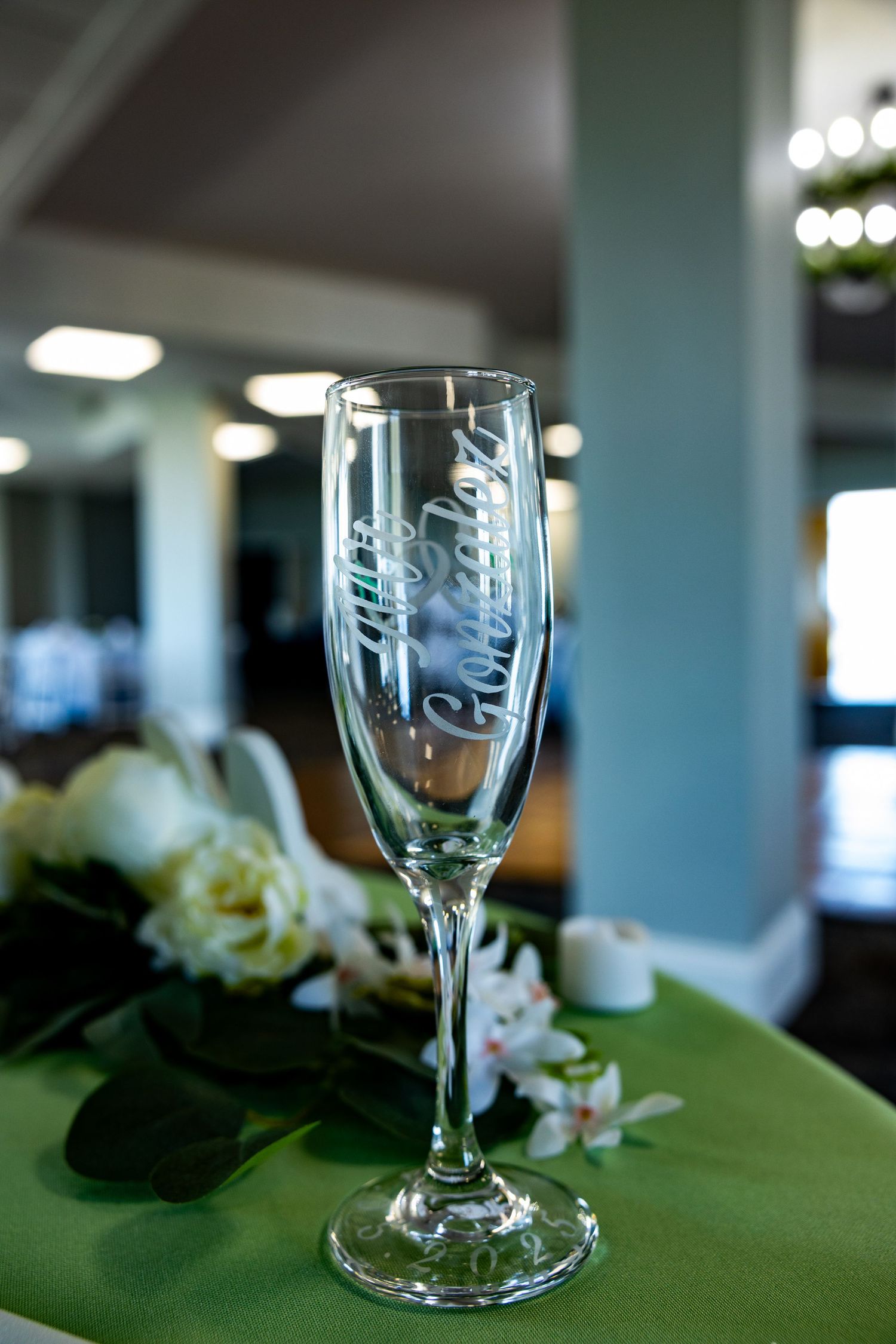 An engraved champagne flute sits on a green tablecloth with white flowers in the background.