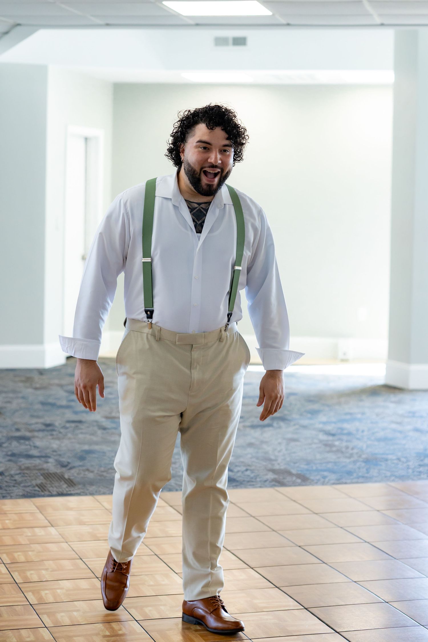 A person in a white shirt and khaki suspenders walks playfully across a tiled floor in a bright indoor space.