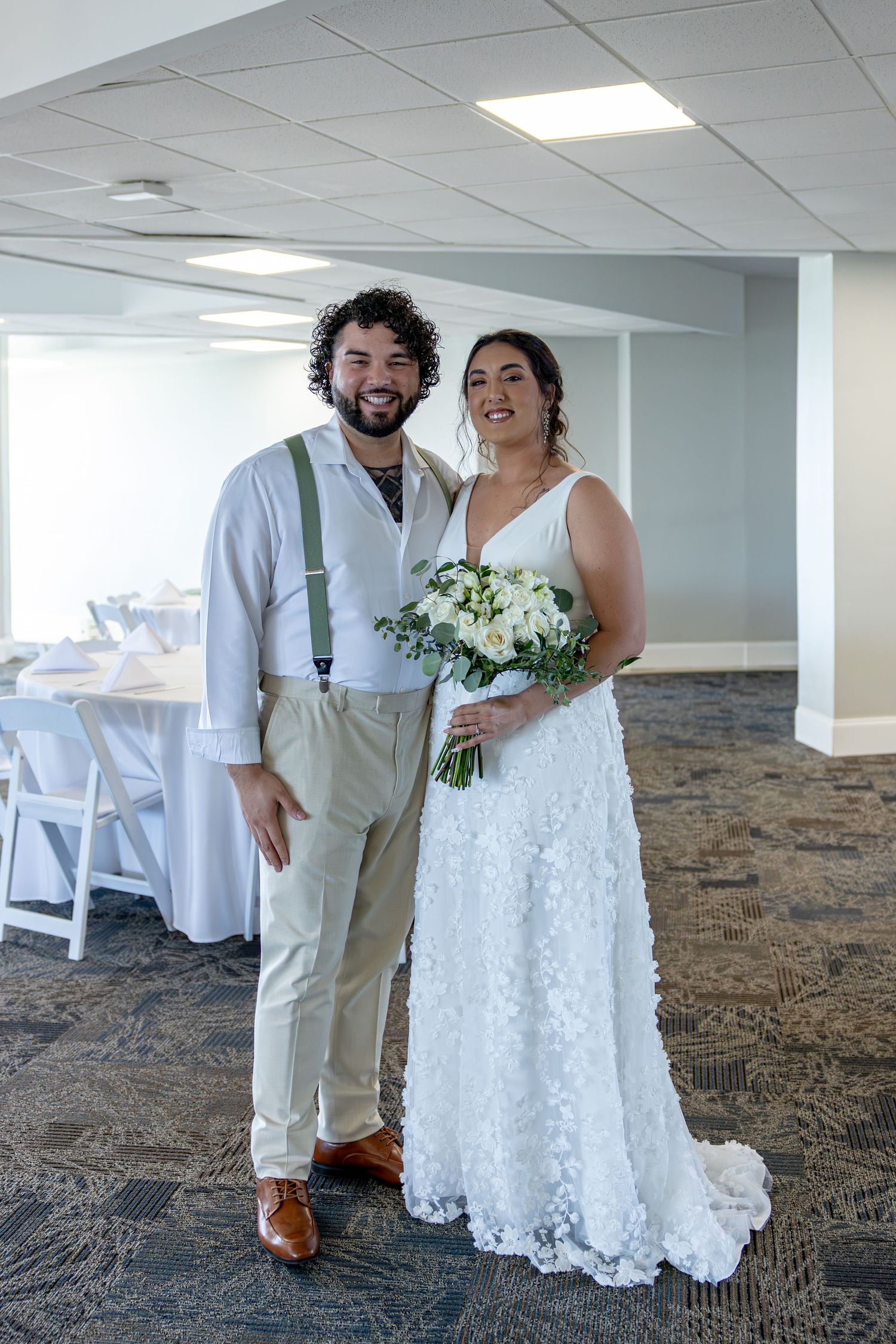 A couple in wedding attire stands together in an indoor venue, wearing white dress and khaki pants with suspenders.