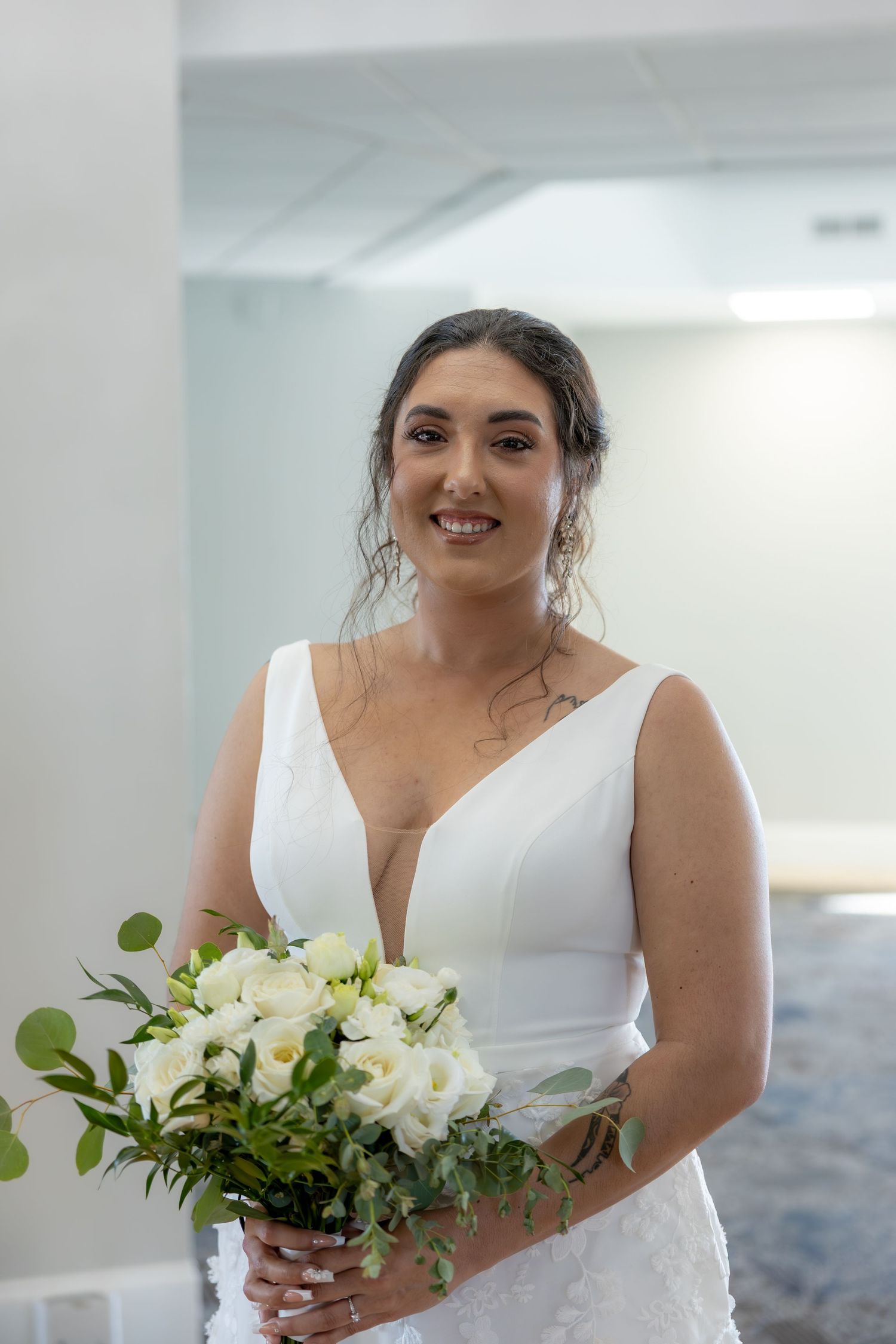 Bride in a white V-neck wedding dress holds a white and green floral bouquet while posing in a bright room.