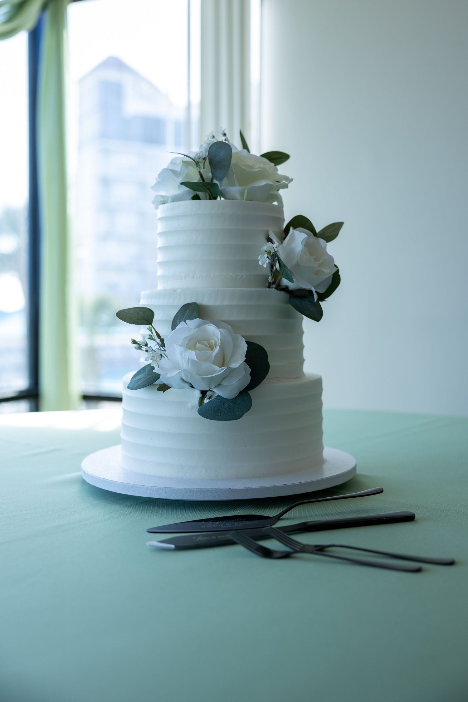 White wedding cake with floral decorations set on a light colored table with serving utensils.