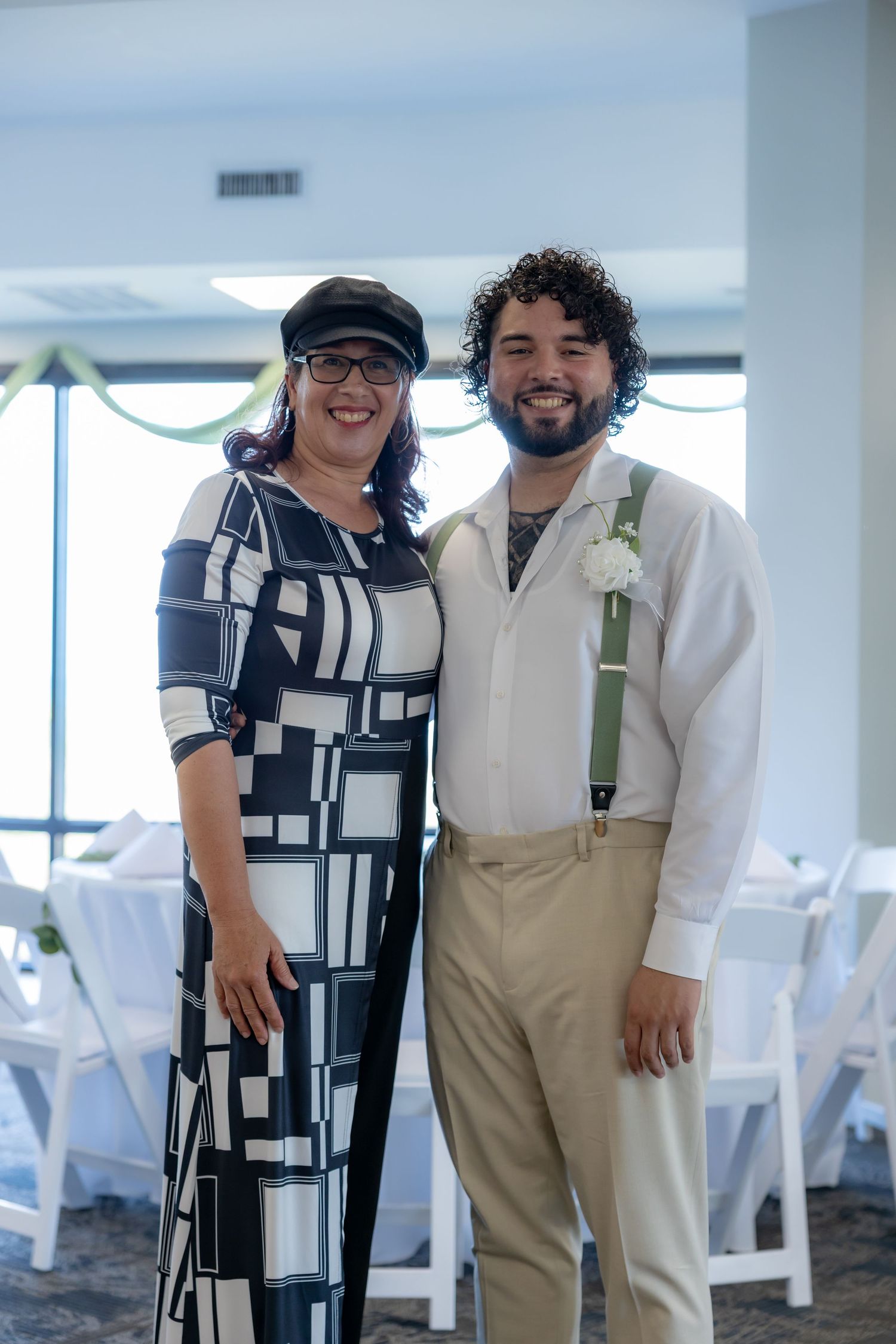 Two people smile and pose together at an indoor event space with white chairs in the background.