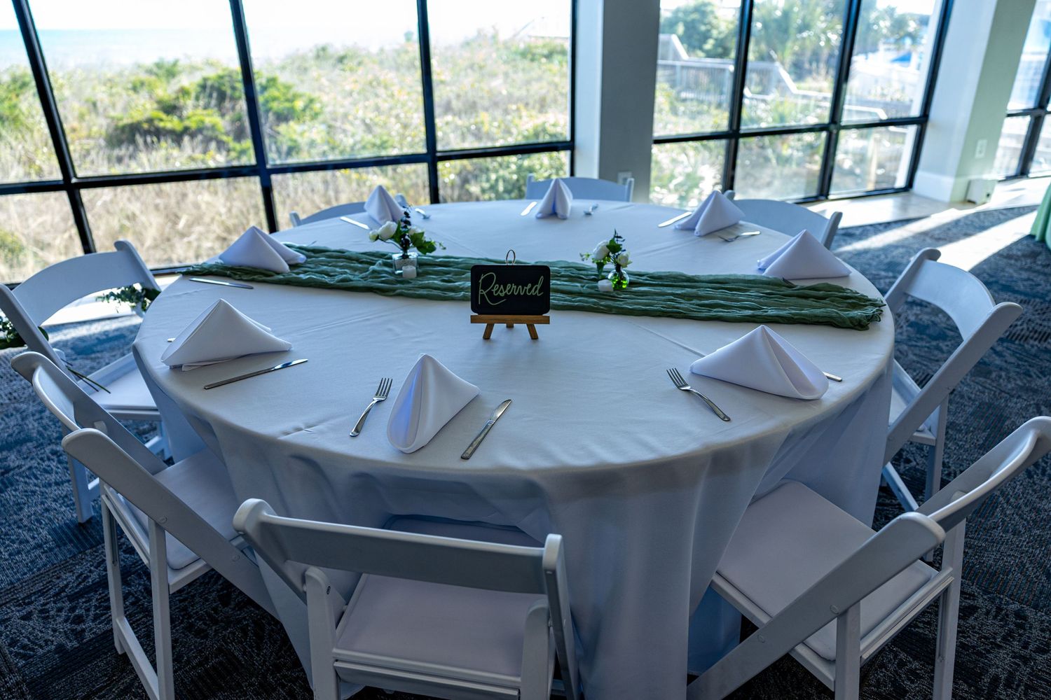 A round white table set for a wedding reception with place settings and a centerpiece near large windows.
