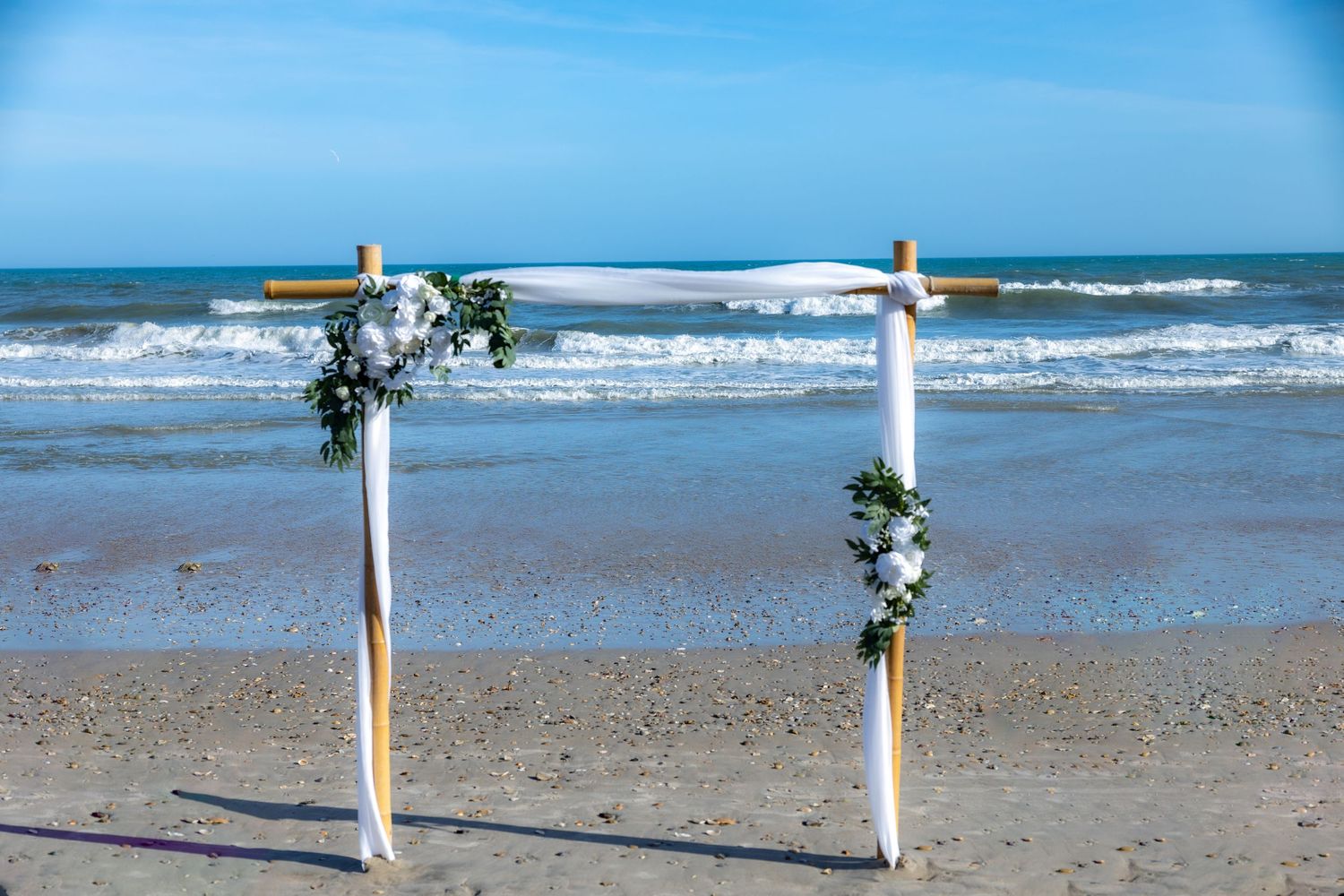 Beachfront wedding ceremony setup with white posts and floral arrangements along the sandy shoreline.