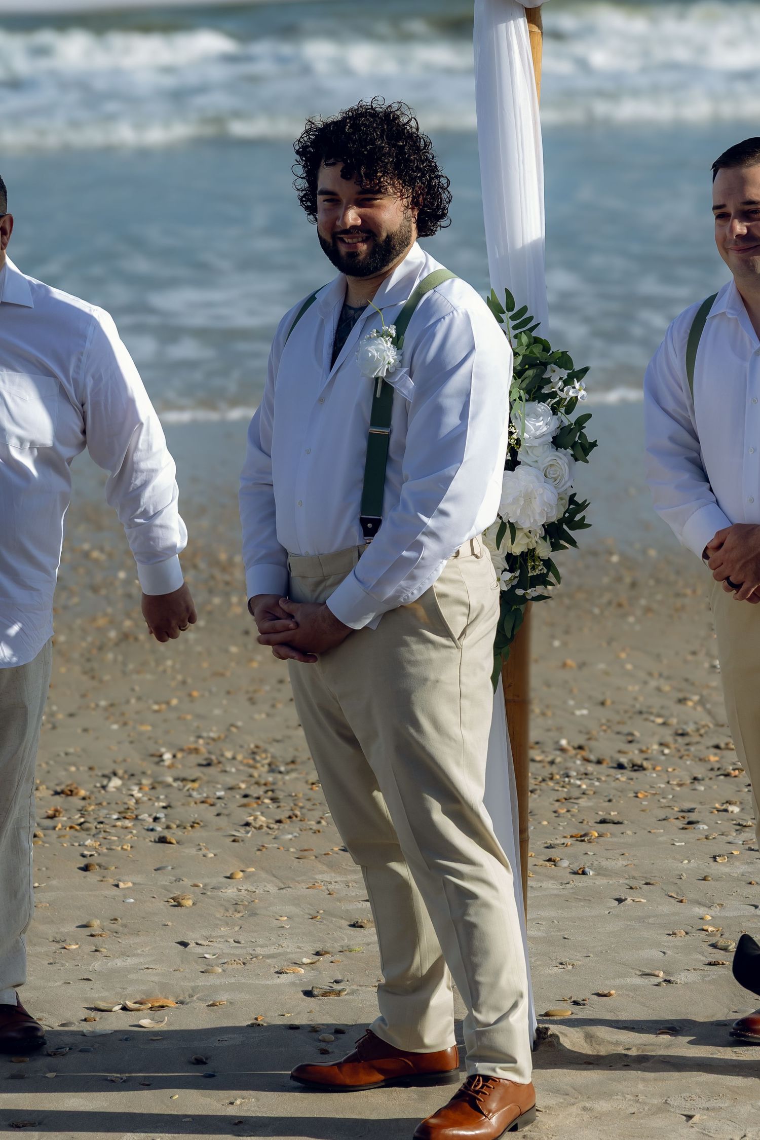 Wedding party members stand on beach in white shirts and khaki pants near ocean.