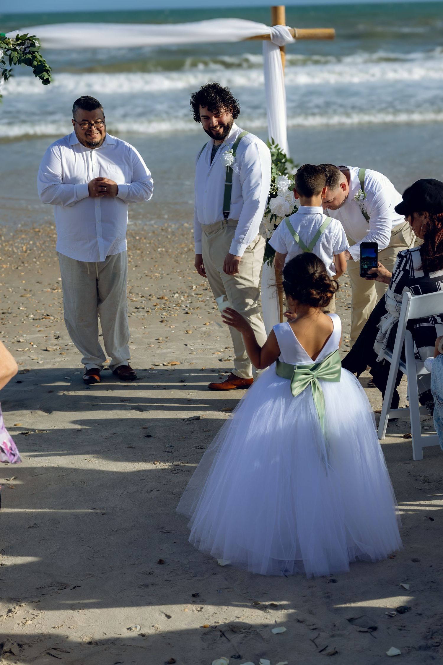 Beach wedding ceremony setup with attendees gathered on sandy shore near ocean.