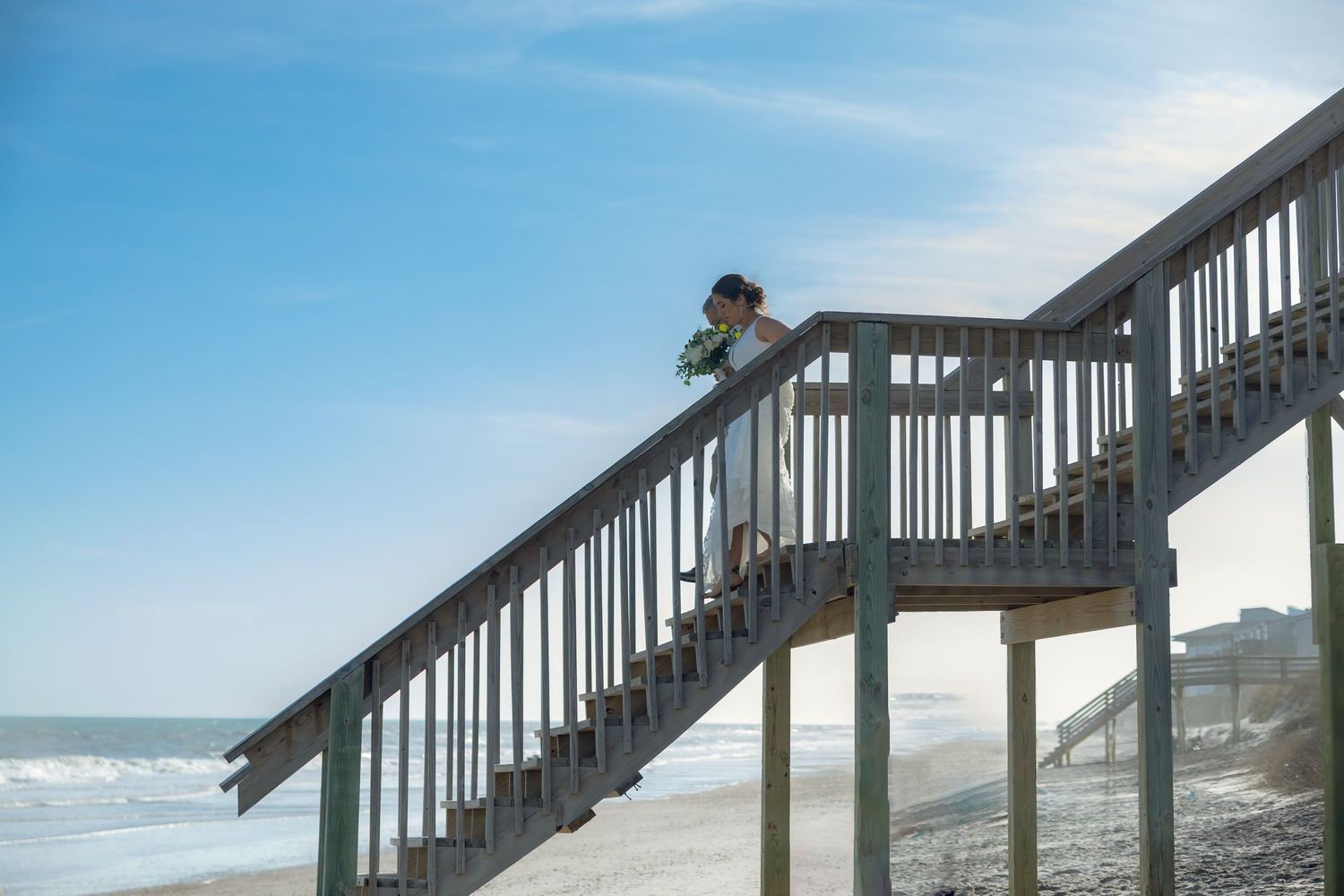 A wooden staircase leads down to a sandy beach with ocean views on a sunny day.