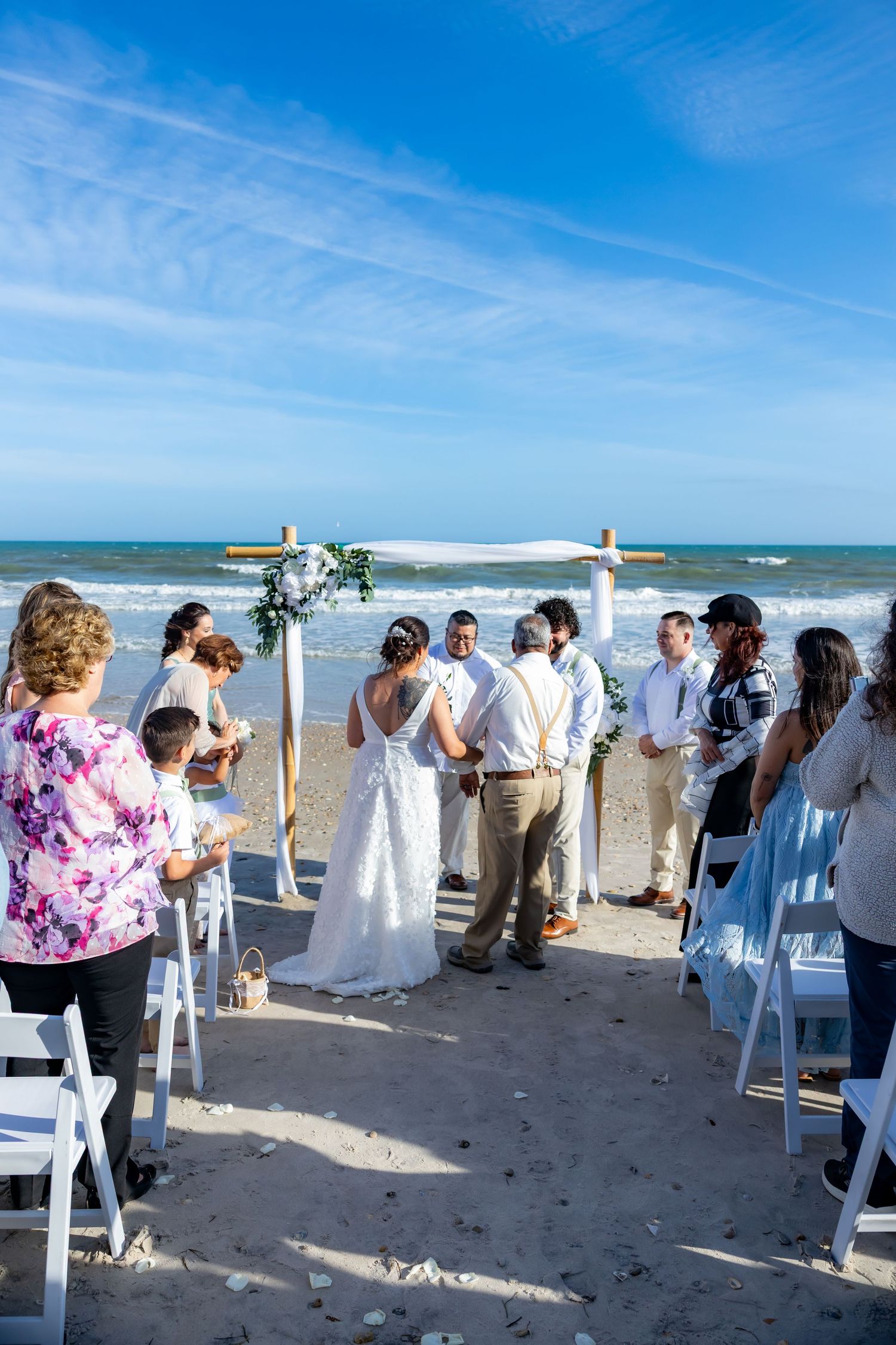 Beach wedding ceremony under a bamboo arch with guests seated on white chairs by the ocean.