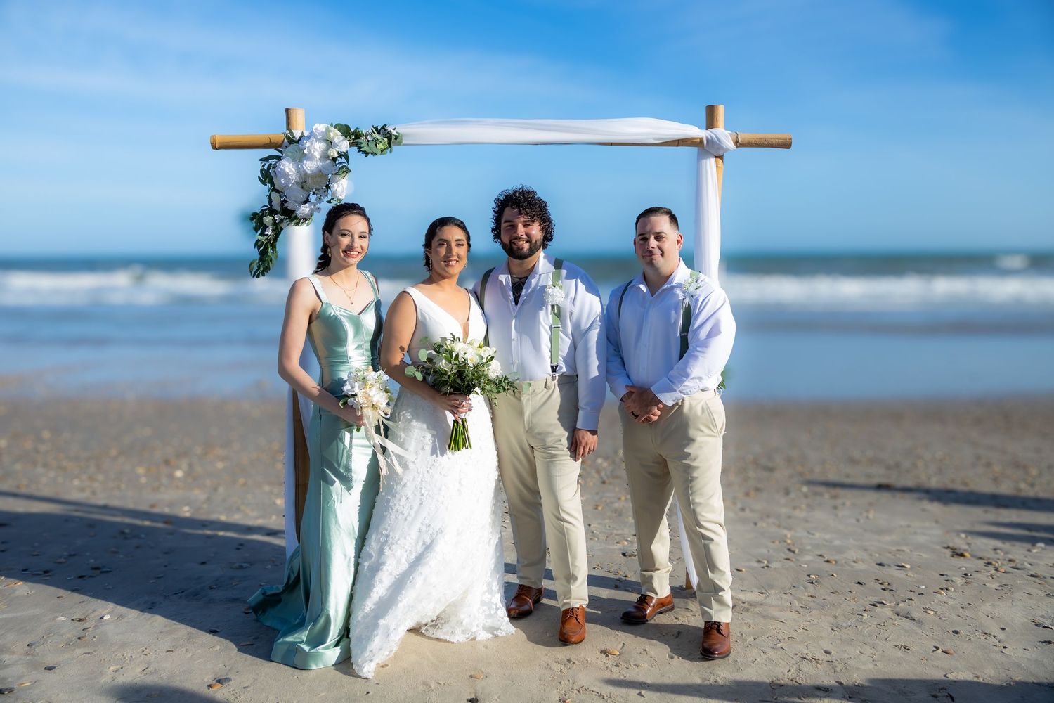 Wedding party poses together on beach with wooden cross and ocean backdrop.