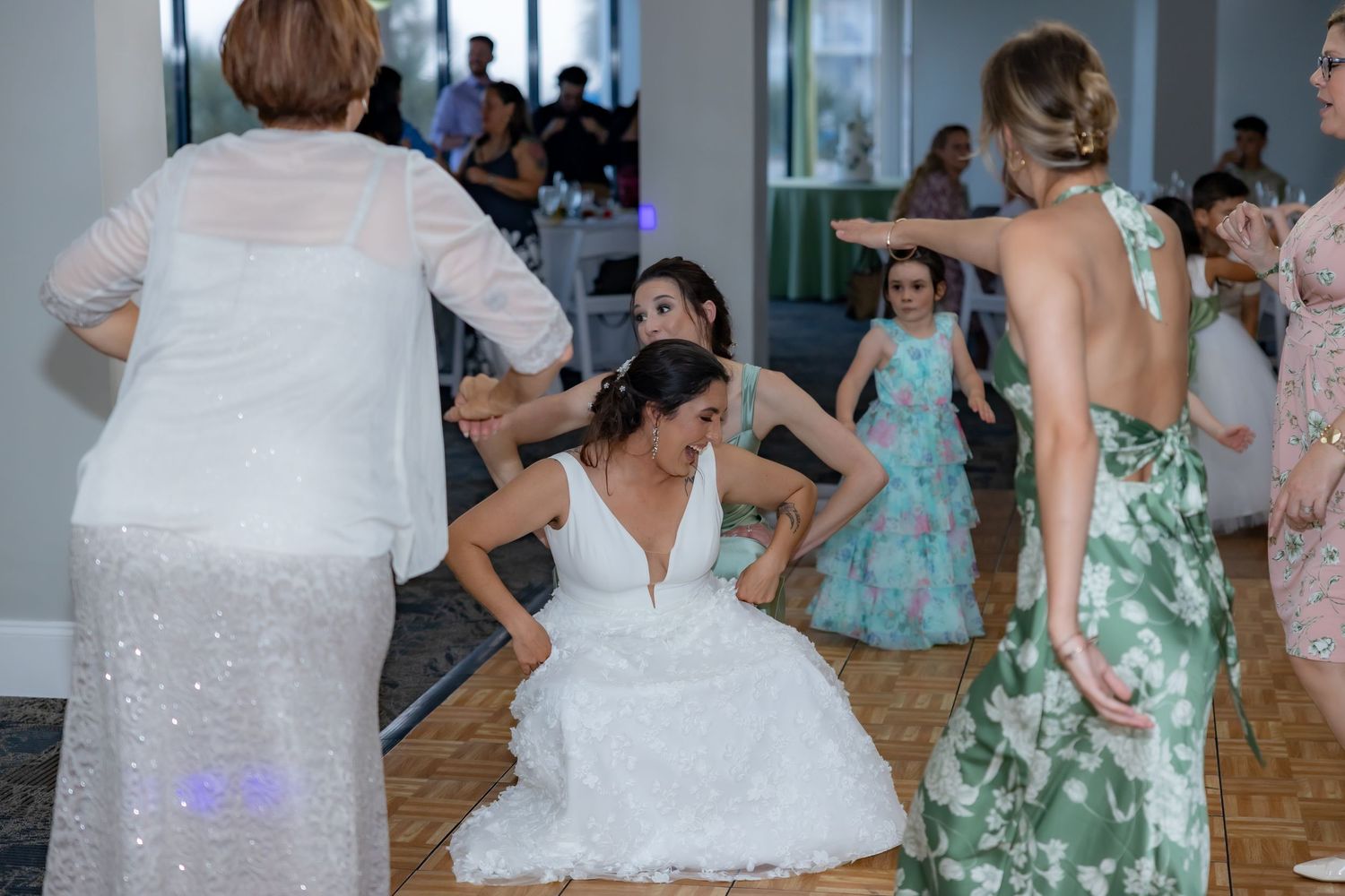 Wedding party guests dance and celebrate on a wooden dance floor at an indoor reception venue.
